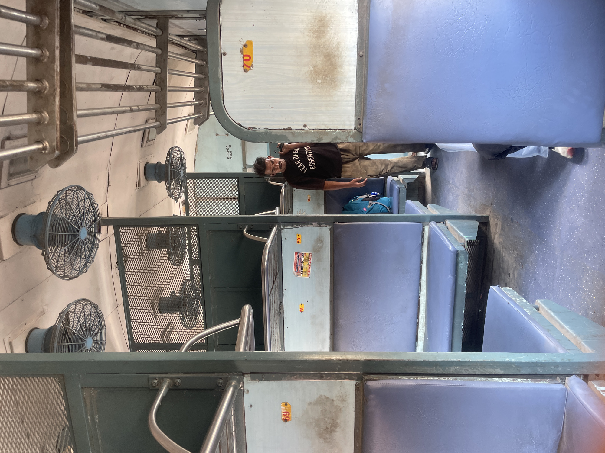 The photograph shows the interior of a passenger train carriage. The carriage features tiered seating, with blue upholstered seats visible. The walls and ceiling are painted a pale, possibly off-white, color, and metal grates and supports are present throughout. Several circular fans are mounted on the ceiling.
A man wearing a black t-shirt with white text and tan pants is standing in the aisle. He has dark hair and a mustache and is holding a dark object over his shoulder. A blue backpack is visible on a seat behind him. The floor of the carriage appears to be a concrete or tiled surface with some debris.
Visible on the side of the carriage are yellow and orange numbered tags, possibly indicating seat numbers. One tag reads "69", another "78", and a third "79", and a fourth "70". There is also a sticker with text that appears to be in a non-English script, with numbers and possibly an image. A sign on the wall reads "GS J43456 TO SEAT - 90".
The scene is likely within a railway station or moving train in Pathankot, India, given the style of the carriage and the presence of typical Indian railway infrastructure.
