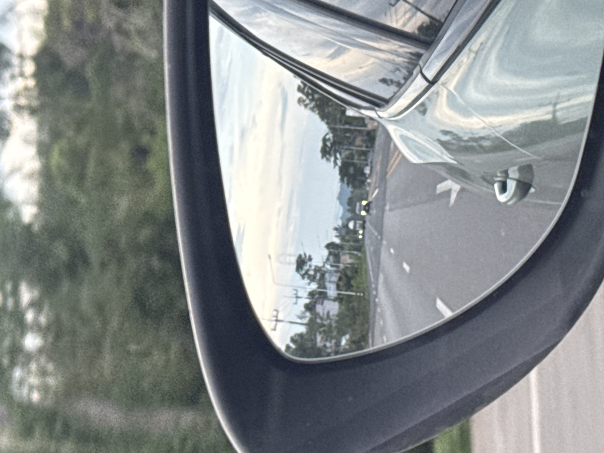 The image is a reflection in a car's side mirror, showing a road in Don Sak, Thailand. The road is paved with white lane markings and appears to be a two-lane highway. On the right side of the road, there are trees and buildings, along with utility poles. In the distance, there are two vehicles, one of which is a truck. The sky is overcast with some clouds visible. The car's body is visible in the foreground, reflecting the sky and trees. The overall scene suggests a daytime drive on a rural or suburban road.