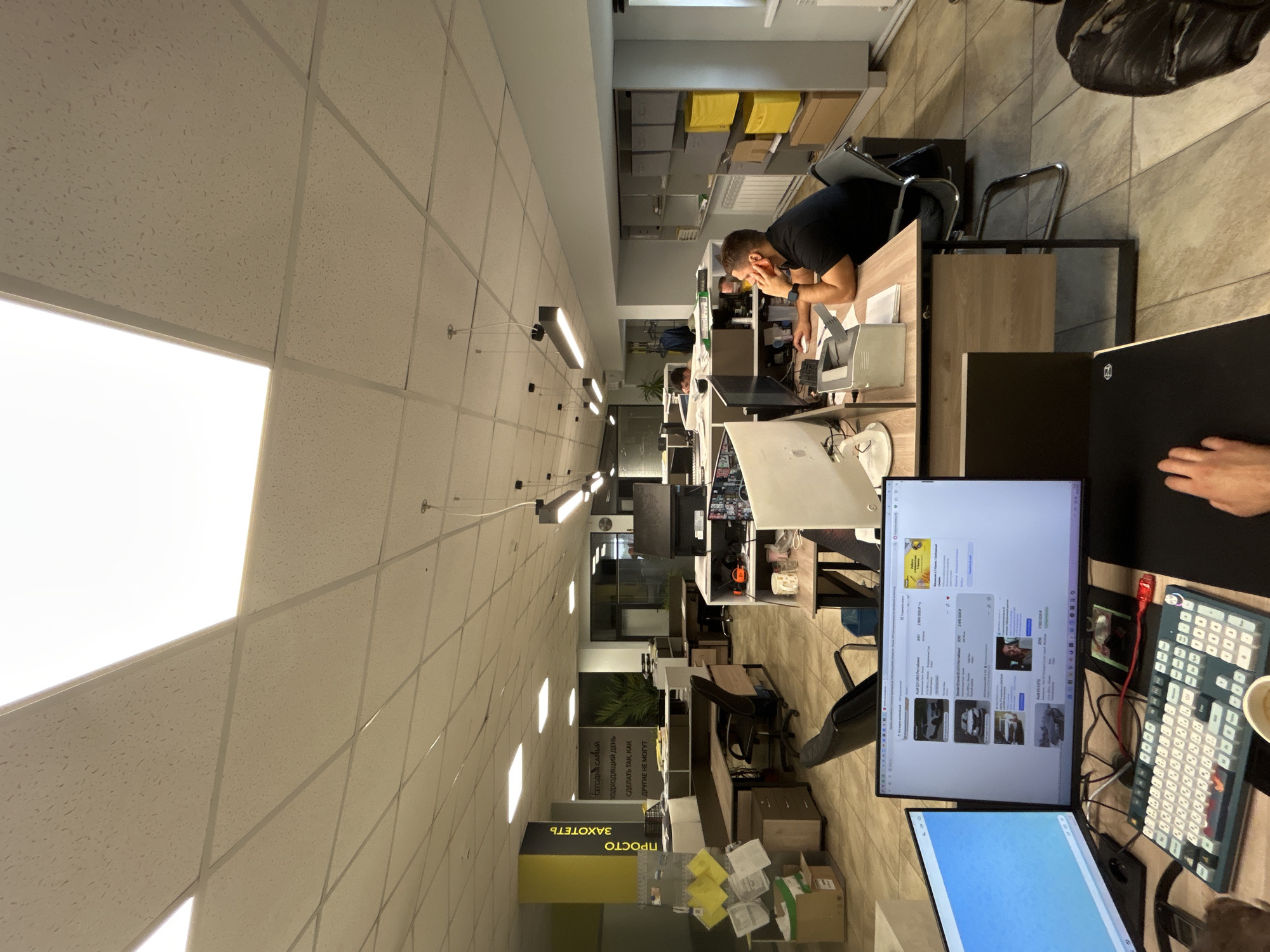 The image depicts an office environment, likely in Votkinsk, Russia based on the Cyrillic signage ("ПРОСТО ЗАХОТЕТЬ," meaning "Just Want") visible at the bottom. The office features rows of desks, each equipped with computer monitors, keyboards, and other office supplies. One individual is visible, seated at a desk and working on a computer. The environment appears to be a modern workplace with a focus on technology. Overhead, the ceiling has fluorescent lighting panels. The floor is tiled. Storage units with boxes are visible on the upper part of the image near the first person. The image has been rotated to show the details better.
