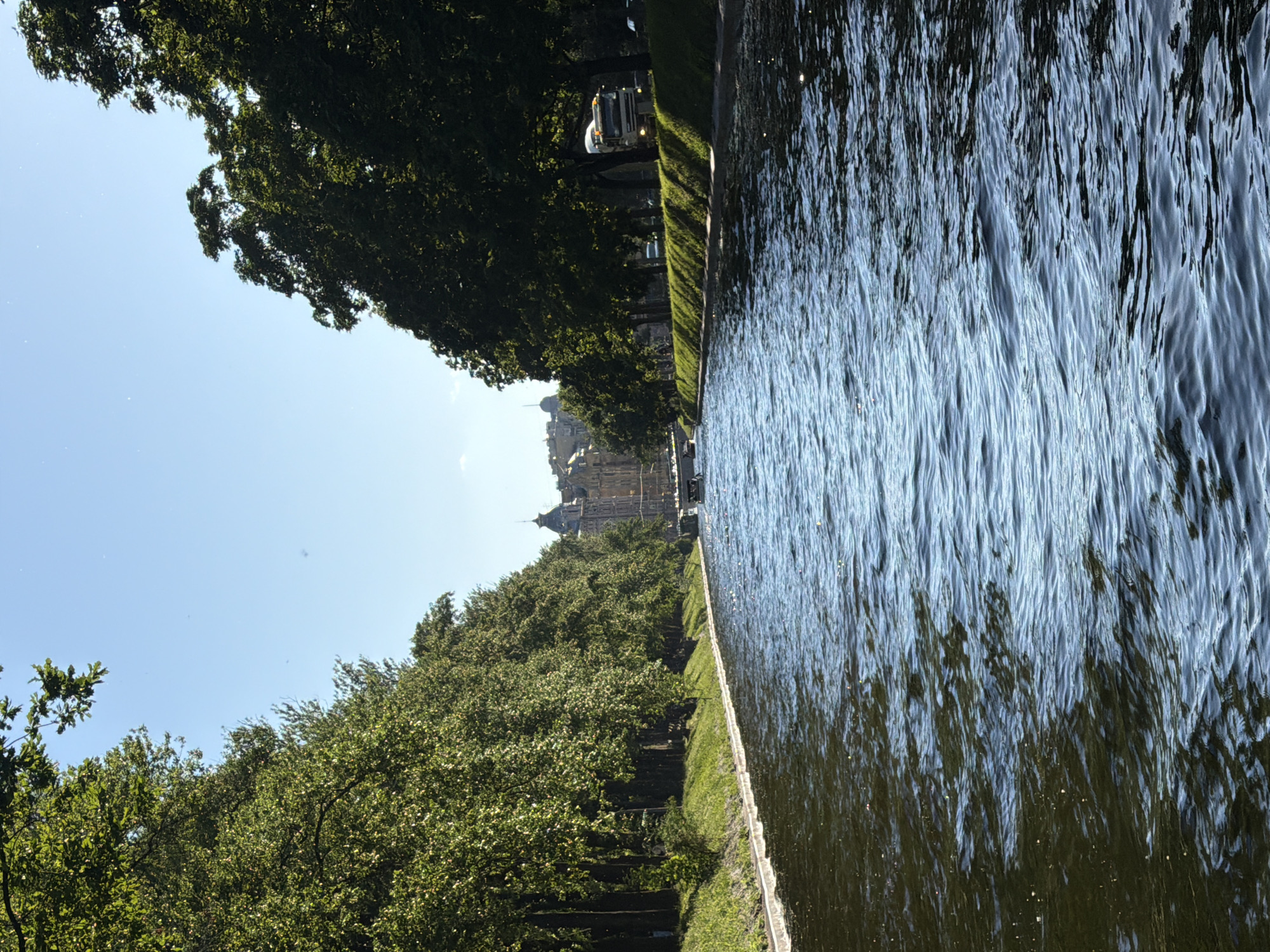 The image depicts a serene canal lined with lush green trees, a picturesque scene in Saint Petersburg, Russia. The canal's water, reflecting the bright sky and surrounding foliage, creates a rippling, dynamic surface. On the left bank, a dense row of trees with vibrant green leaves forms a natural canopy, with glimpses of buildings and spires of a historical castle or palace peeking through the foliage in the distance. The architecture suggests a grand, classical style, typical of Saint Petersburg's historic districts. A street cleaner vehicle is visible on the right side of the canal, moving along the path next to the trees, indicating some urban activity amidst the natural beauty. The clear blue sky suggests it is daytime, likely a sunny day with pleasant weather, ideal for exploring the city's waterways and landmarks. The overall mood is one of tranquility and timeless elegance, a common characteristic of Saint Petersburg's iconic canals and grand architecture. No visible text is present in the image.