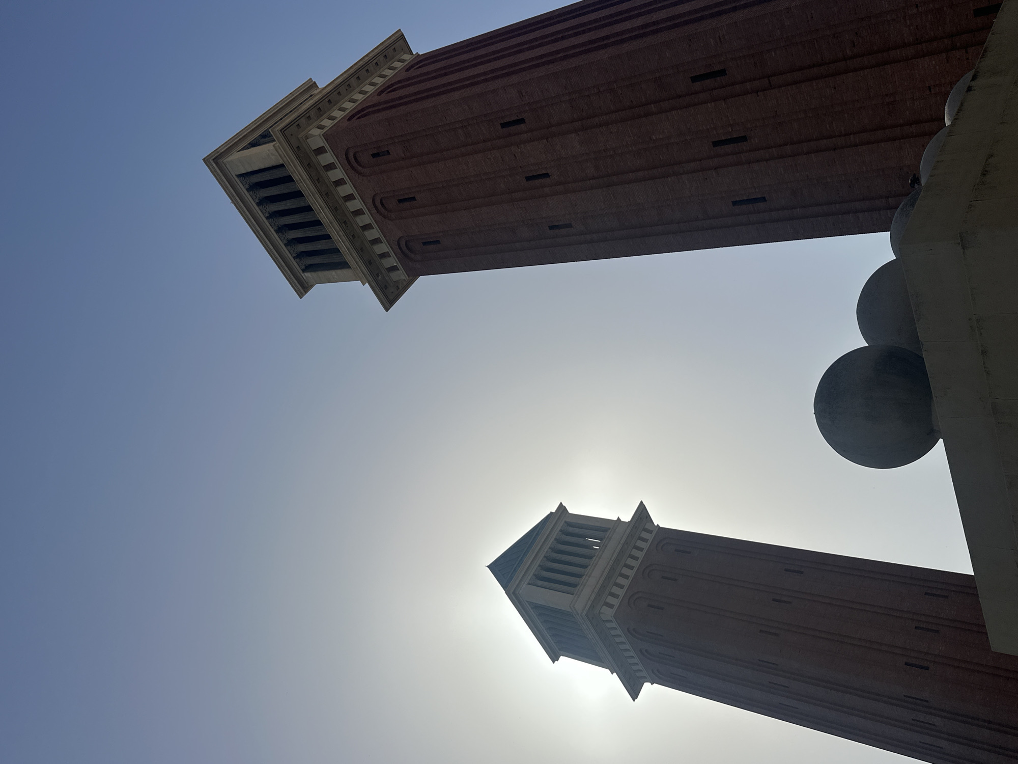 The image shows a low-angle view of two tall, brick towers with decorative tops against a bright, hazy sky. The towers appear to be part of a larger architectural complex. The top portion of the right tower is partially obscured by a large, concrete structure with spherical elements. The sun is a bright white disc behind the towers, suggesting it is either sunrise or sunset, or a very bright, overcast day. The overall impression is one of grandeur and architectural scale. The image appears to have been taken in Barcelona, Spain.