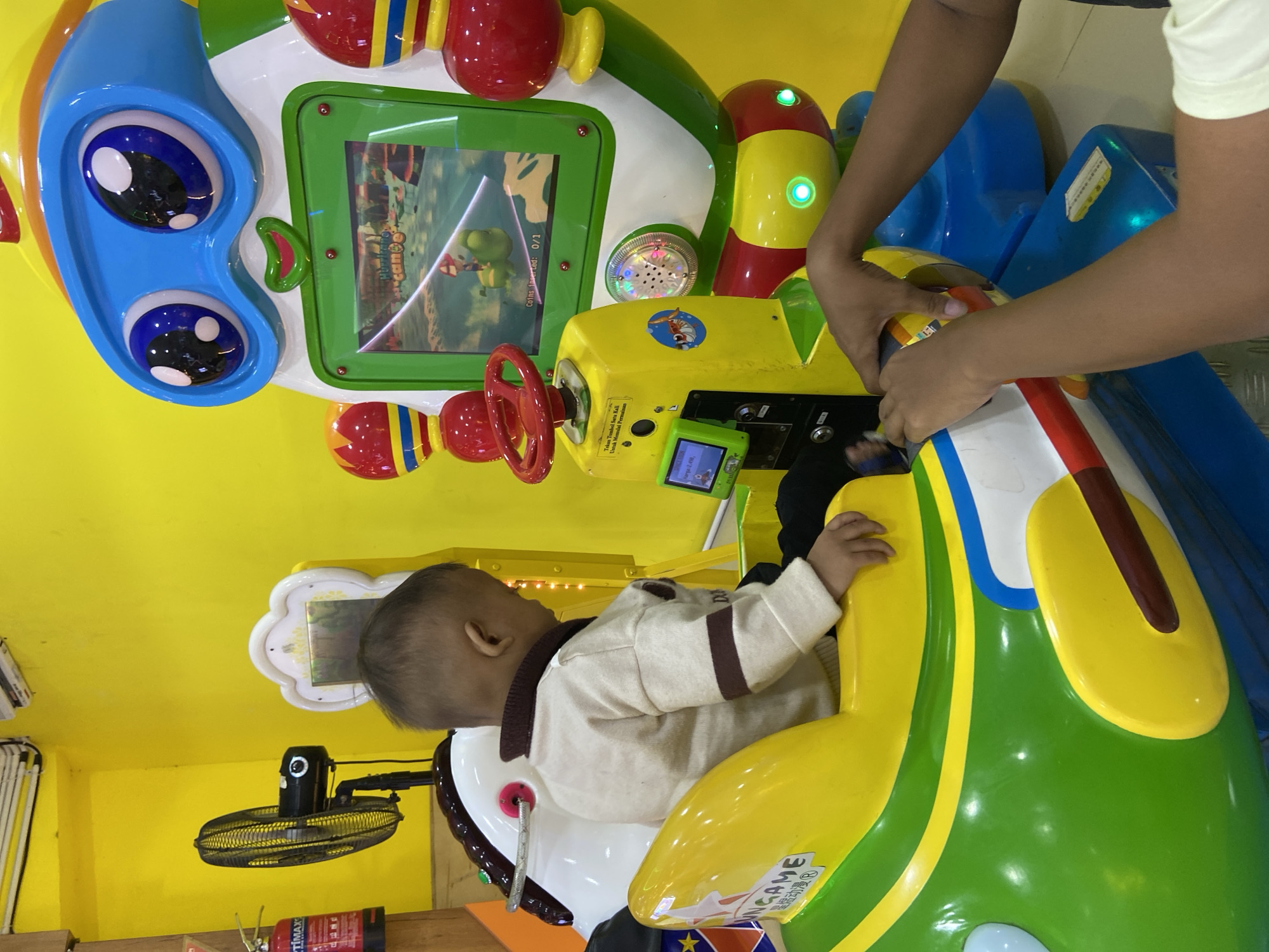 A child is seated in a colorful arcade ride, shaped like an airplane, in a room with bright yellow walls. The child wears a light-colored sweater with dark trim. To the left of the child is a black oscillating fan. To the right, a large, cartoonish robot-like structure with blue goggles and red and yellow accents houses a video game screen displaying cartoon characters. The screen shows "Coins Inserted: 0/1". A red steering wheel is attached to a yellow control panel in front of the child. The floor appears to be a dark tiled surface with metallic accents. The overall environment suggests an indoor amusement or entertainment venue. The image appears to have been taken in Kecamatan Pati, Indonesia, as indicated by text visible on the side of the arcade ride.
