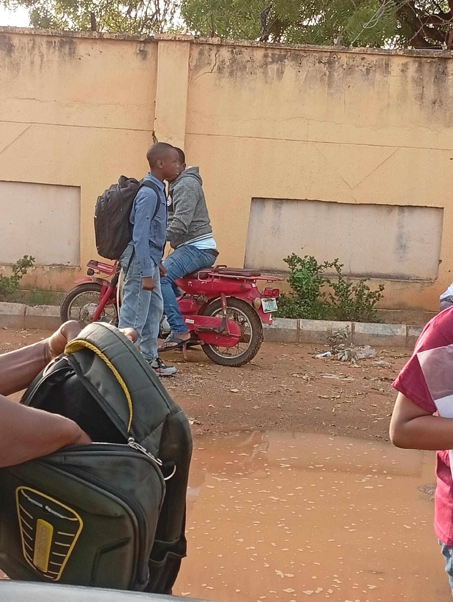 The image depicts two young men on a red motorcycle parked on a dirt road next to a yellow wall in Barden Yana, Nigeria. One man is wearing a blue shirt, jeans, sneakers, and a backpack while standing next to the motorcycle. The other man is wearing a grey hoodie, jeans, and sandals while sitting on the motorcycle. In the foreground, there is a green and black backpack, partially open. There is a puddle on the dirt road. A person wearing a maroon shirt stands to the right of the image.