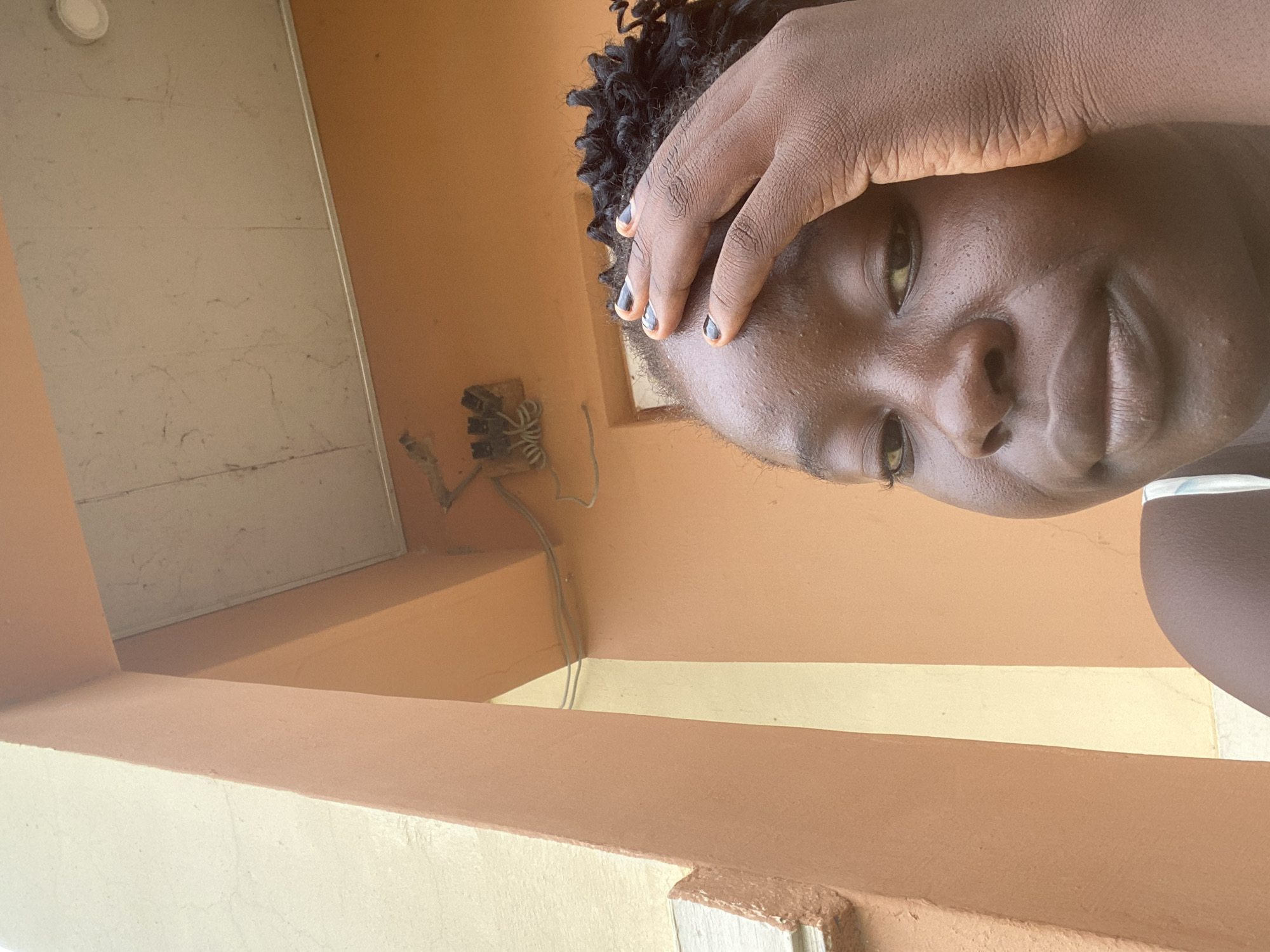 This is a close-up, low-angle, portrait-oriented, outdoor photo of a young Black person. Their face is the primary subject, turned towards the camera with their left hand pressed to their forehead. Their eyes are open and looking at the camera, with a neutral or slightly pensive expression. Their skin is dark, and their hair is styled in short, dark, curly locks. They have painted nails with a design of black tips and a copper-orange base.
The background is out of focus and consists of architectural elements. To the left, a light-colored surface with horizontal lines and a circular light fixture can be seen. To the right and below, there are painted walls in shades of orange and yellow. Wires and a small, grey electrical junction box are visible on the orange wall in the mid-ground. The lighting suggests it is daytime, possibly sunny, judging by the bright illumination and shadows.
The scene appears to be an outdoor space with some visible infrastructure, perhaps a porch, balcony, or an exterior wall of a building. The overall impression is a candid moment captured outdoors in Bulamare, Nigeria.