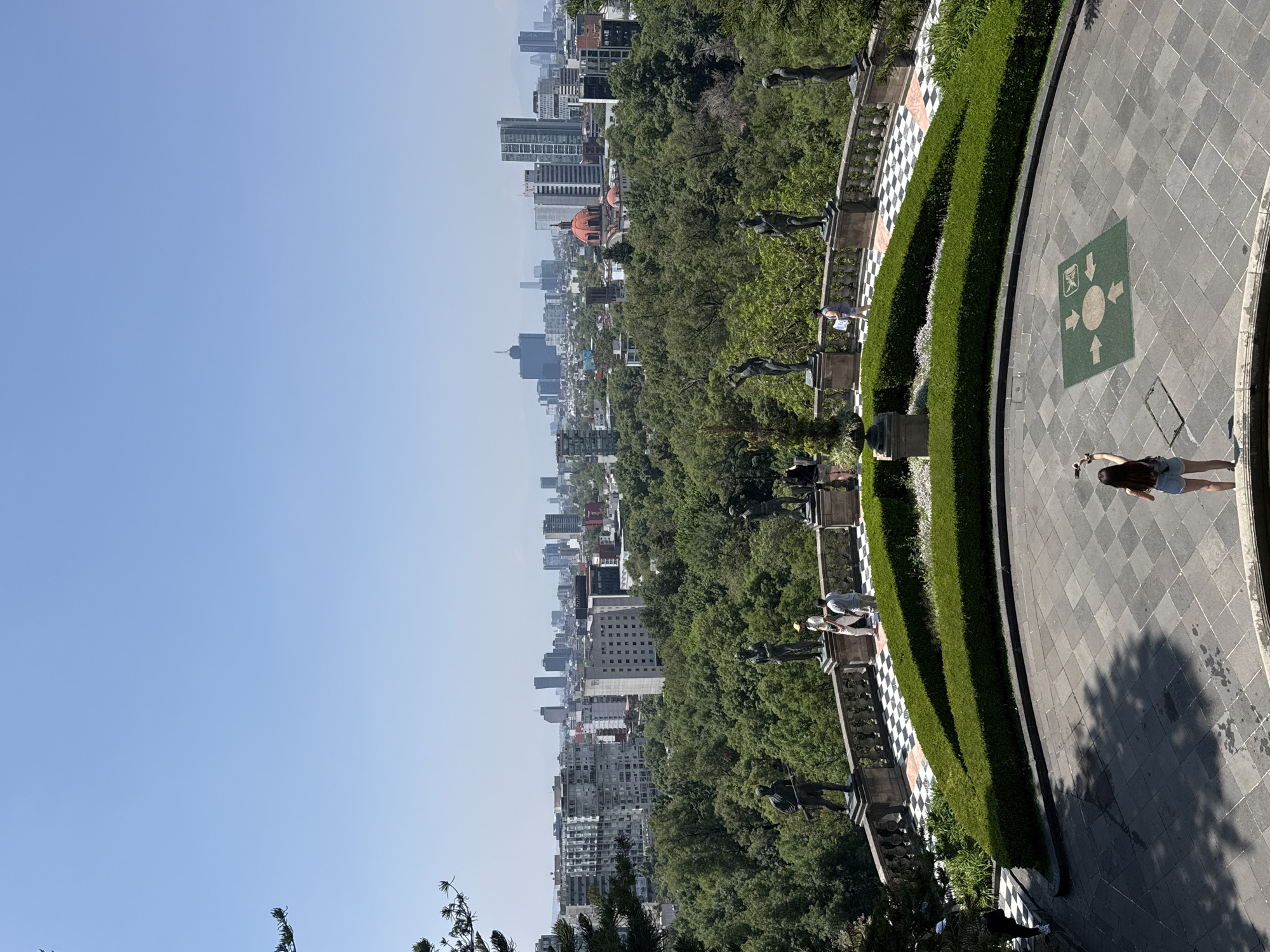 The image shows a partial view of Mexico City, Mexico, taken from a high vantage point. The foreground depicts a stone walkway or plaza, paved in a checkered pattern of grey tiles, with a patch of greenery and a black-and-white checkered wall bordering it. A person is standing on the walkway, facing away from the camera, holding what appears to be a cell phone.
Beyond the walkway, there is a dense green area, which seems to be a large park or forest. Along the edge of the park, there are statues on pedestals. In the distance, there is a view of Mexico City's skyline, featuring a mix of high-rise buildings and other structures. The sky is a clear, pale blue.