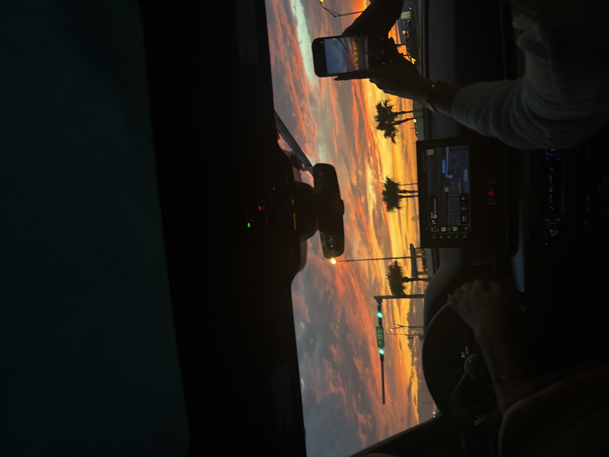 The image captures a sunset scene from inside a car, possibly in Miami, United States of America, as indicated by the presence of palm trees and the style of streetlights. The sky is filled with vibrant colors of orange, pink, and yellow, with clouds creating a textured effect. In the foreground, a person is driving, with their hands on the steering wheel. To the right, another person is holding up a smartphone, taking a picture of the sunset. The car's dashboard displays a navigation screen. Streetlights and traffic signals are visible in the background. The overall impression is a moment of enjoying a scenic view while traveling.