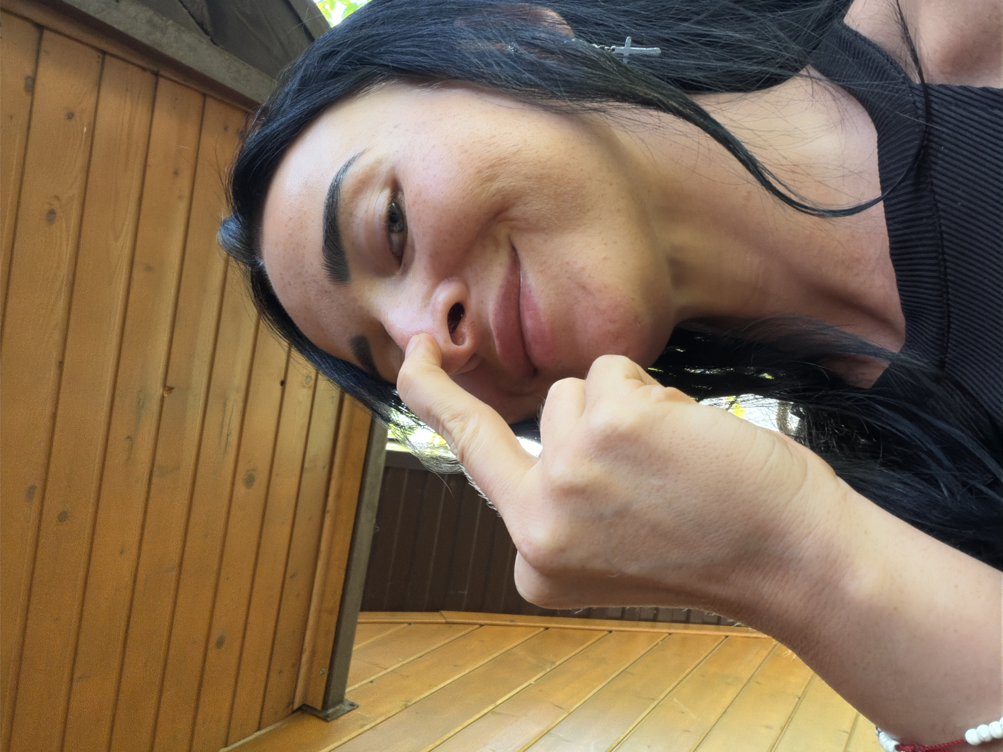 A close-up, low-angle selfie shows a person with long, dark hair. The person is tilting their head and looking towards the camera with a playful smirk, touching their nose with their index finger. Their expression suggests amusement and a lighthearted mood. They are wearing a black, ribbed top. The background consists of wooden panels, likely part of an outdoor structure such as a balcony or porch. The wood appears weathered and is oriented vertically on the left side of the image and horizontally on the bottom. The lighting suggests it is daytime, with natural light filtering in from the top left, casting subtle shadows. The presence of wood and the overall natural lighting imply an outdoor setting in a temperate or warm climate. The location context of Sochi, Russia, aligns with these observations. No text is visible in the image.