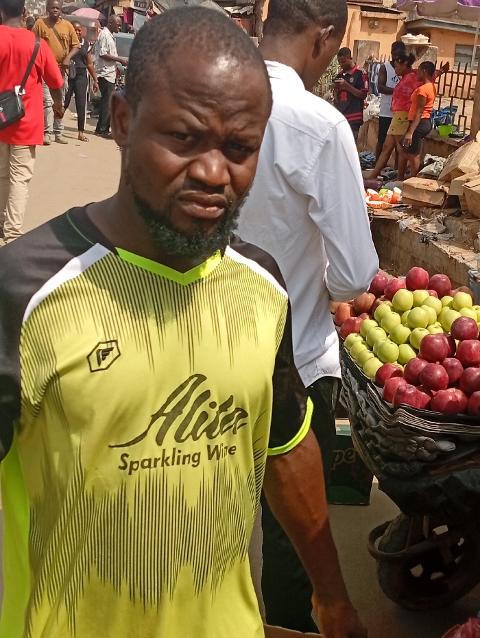 A close-up captures a stern-faced man wearing a yellow and black t-shirt with "Alita Sparkling Wine" printed on it. He stands near a cart overflowing with red and green apples. Several individuals are blurred in the background, suggesting a bustling marketplace. The scene is sunlit, with a focus on the man's expression and the vibrant fruit.