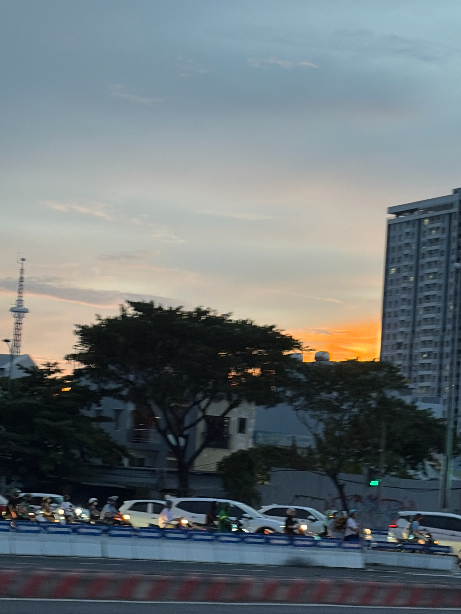 The image depicts a street scene in Da Nang, Vietnam, at dusk. A tall, modern building dominates the right side of the frame, its windows illuminated. To the left, a communication tower stands against a sky painted with hues of pale blue, gray, and a striking orange sunset. In the foreground, a busy road is visible, with several cars and numerous motorcycles carrying passengers. The vehicles are separated by a white and blue barrier. Trees with dark, dense foliage are scattered throughout the scene, some obscuring parts of the buildings and the road. The overall impression is one of urban life transitioning into evening, with traffic and the city skyline set against a dramatic sky.
