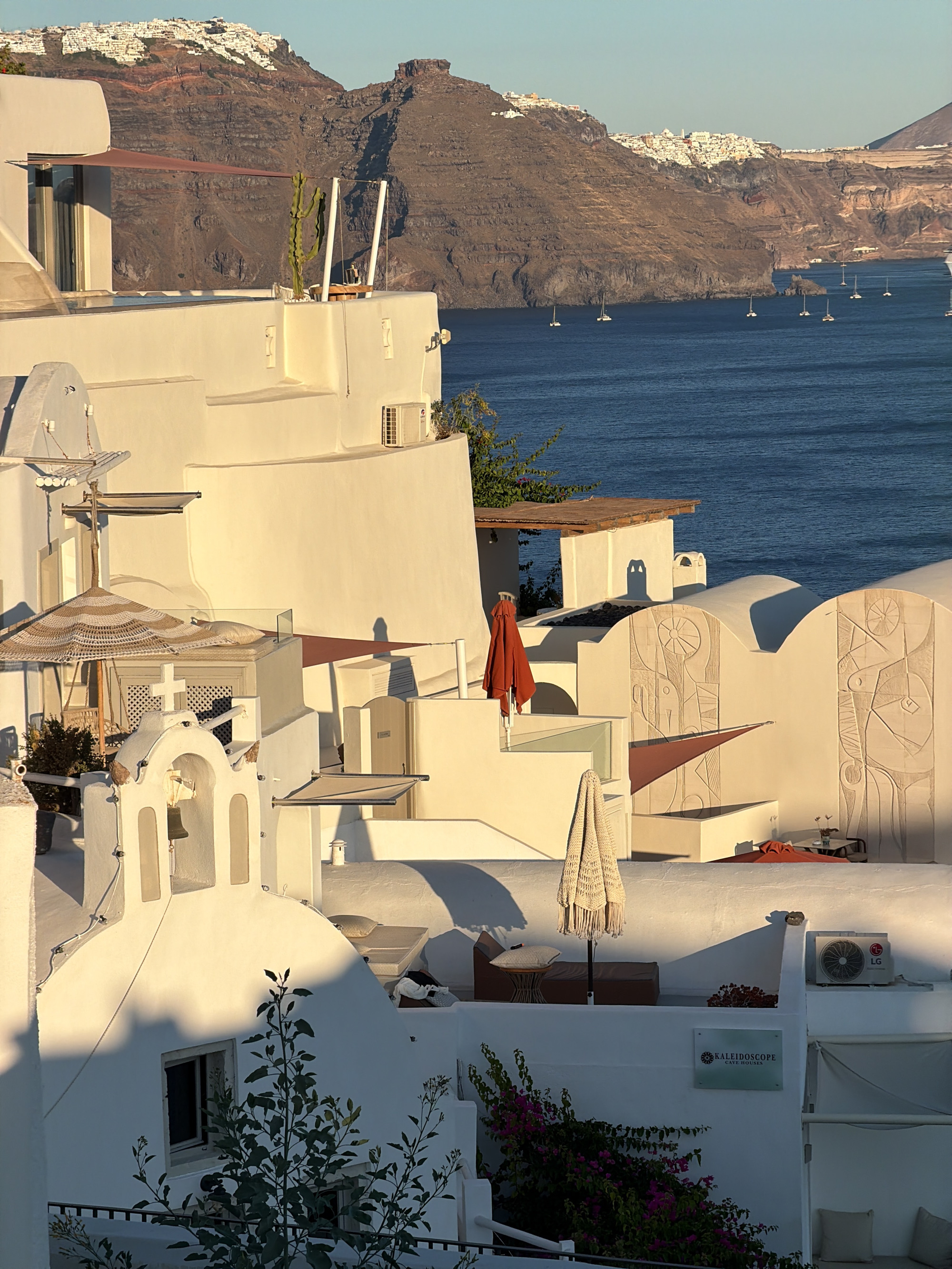 The image depicts a view of a building in Ia, Municipal Unit, Greece, characterized by its whitewashed architecture and terraces overlooking a body of water. The sun is low in the sky, casting long shadows and highlighting the textures of the buildings and the rocky landscape in the background.
In the foreground, a white bell tower with a cross on top is visible. To the left, a portion of a white building with a window and greenery in front is seen. A striped umbrella and a pillow rest on a cushioned seating area.
The midground showcases more of the white architectural style with multiple levels, balconies, and shaded areas. There are several umbrellas, including a red one propped against a wall and a tan crocheted umbrella. A sign for "KALEIDOSCOPE CAVE HOUSES" is mounted on one of the white walls. An air conditioning unit is mounted on the exterior wall. Carved reliefs are visible on some of the curved white walls.
The background features the Aegean Sea with several sailboats. Beyond the sea, a steep, rocky cliff rises, dotted with white buildings that form a village on the clifftop. The sky is a clear, pale blue. The overall scene evokes a serene and picturesque Mediterranean setting.