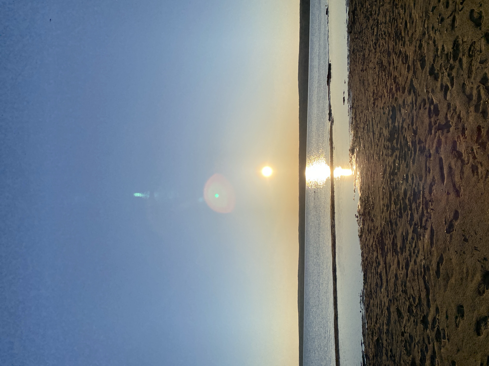The image shows a horizontal view of a shoreline at sunset. The left side of the image depicts a clear blue sky that transitions to a golden hue near the horizon. A bright sun is visible, creating a glare and a lens flare effect with a green spot. To the right of the sun, a body of water extends towards the center of the image, reflecting the sunlight. The rightmost section of the image displays a sandy beach with footprints, suggesting recent human activity. The location is presumed to be a shoreline within the city of Chaikovsky, Russia, given the prompt.