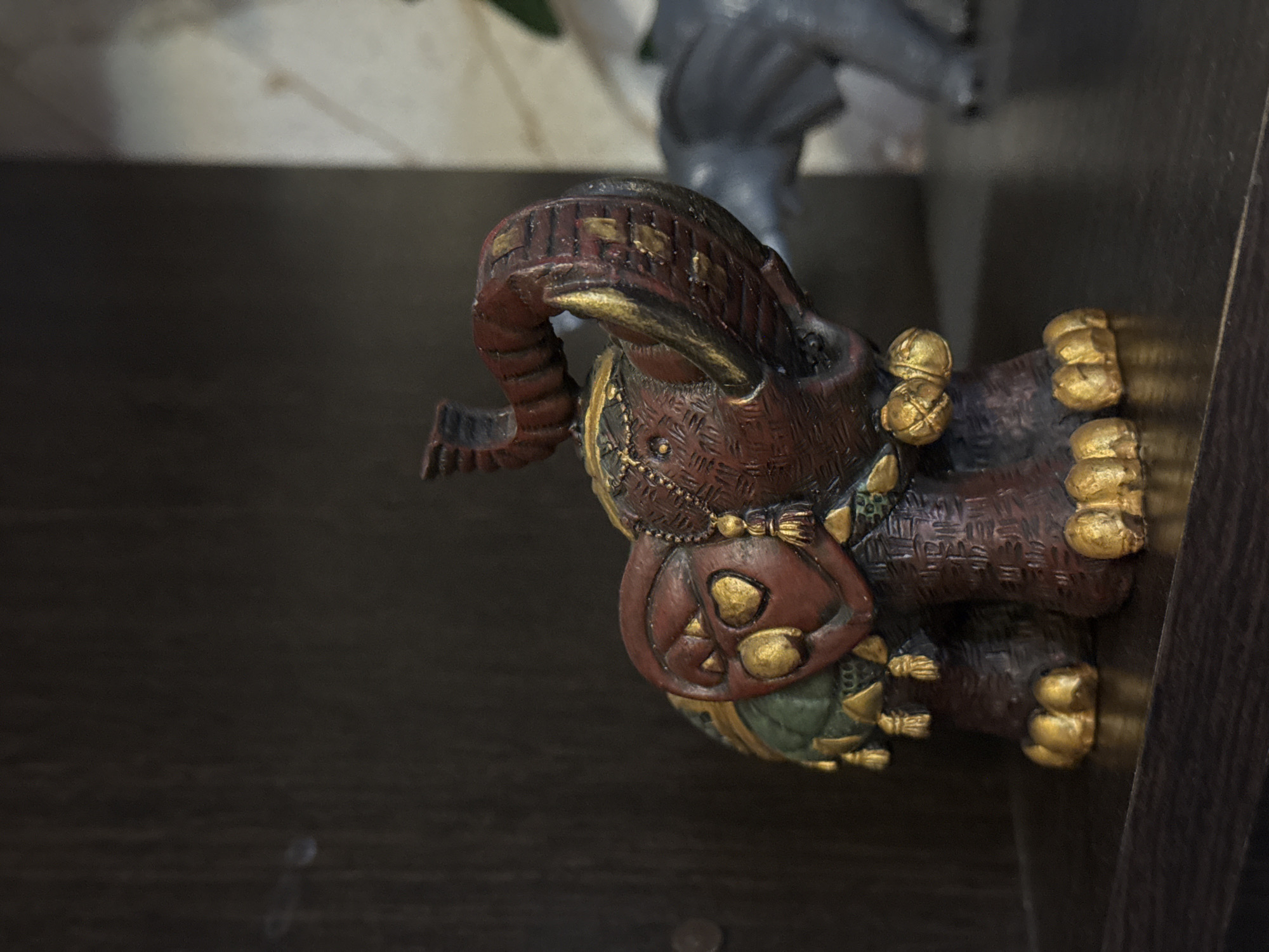 The image features a decorative elephant figurine on a dark brown surface, potentially a shelf or table. The elephant is predominantly reddish-brown with gold accents on its trunk, ears, feet, and saddle. The saddle is round with gold embellishments. The background is out of focus, revealing a light-colored surface and a gray object. The exact location of the image is not definitively determined as Kladovitsy, Russia, and there are no elements in the image to directly indicate the city or country.