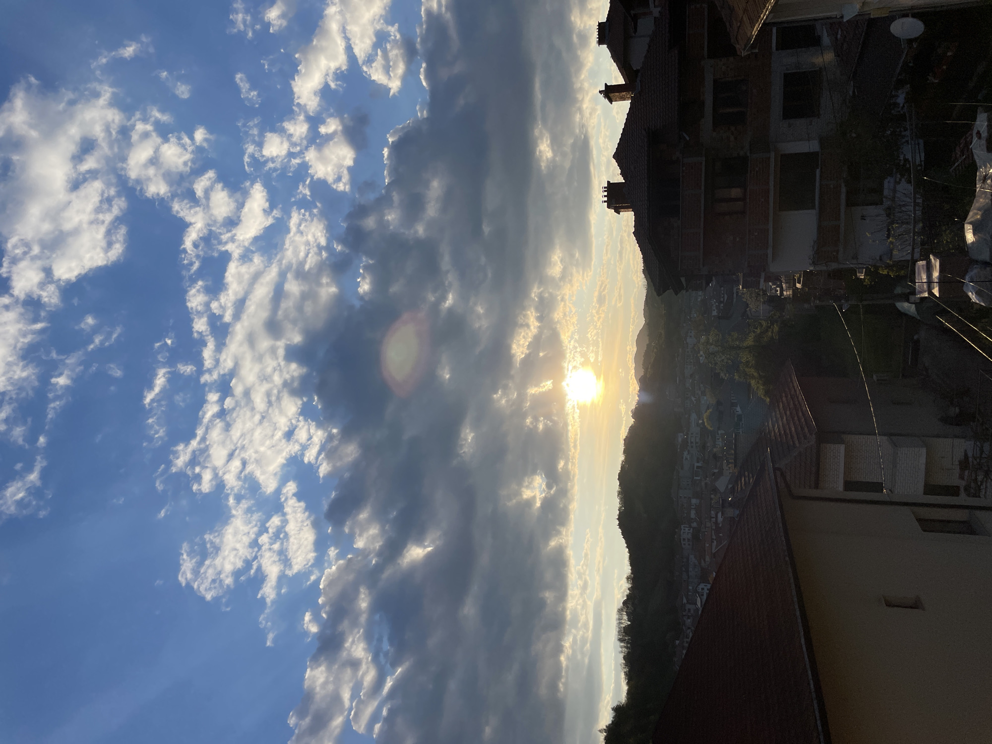 The image shows a view in Zlatograd, Bulgaria, during sunset. In the foreground, sections of buildings with red-tiled roofs and brickwork are visible. The middle ground features a dense area of buildings, indicative of a residential zone, nestled against a backdrop of forested hills. The sun is positioned at the horizon, partially obscured by clouds, creating a bright glare. The sky is filled with scattered cumulus clouds against a blue background on the left side, while darker, stormier clouds cover the center. Overhead power lines are faintly visible between the buildings.