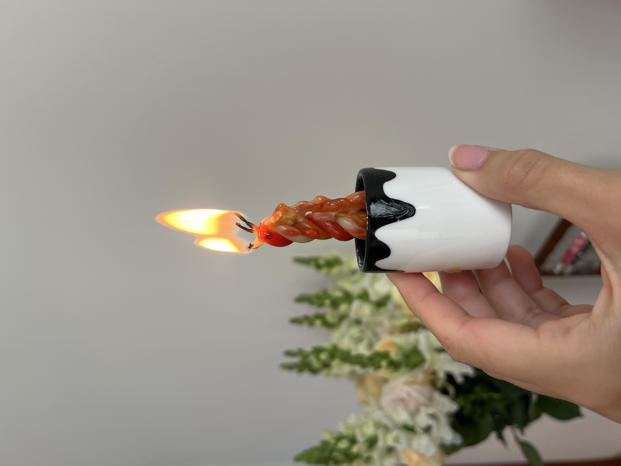 A lit, red and orange twisted candle is held horizontally in a small, white cylindrical holder with a black, scalloped rim. The candle is held by a hand with neatly manicured fingernails. In the background, a blurred arrangement of flowers in shades of white and green sits on a surface. A pale wall is in the distant background. The location is not apparent from the image.