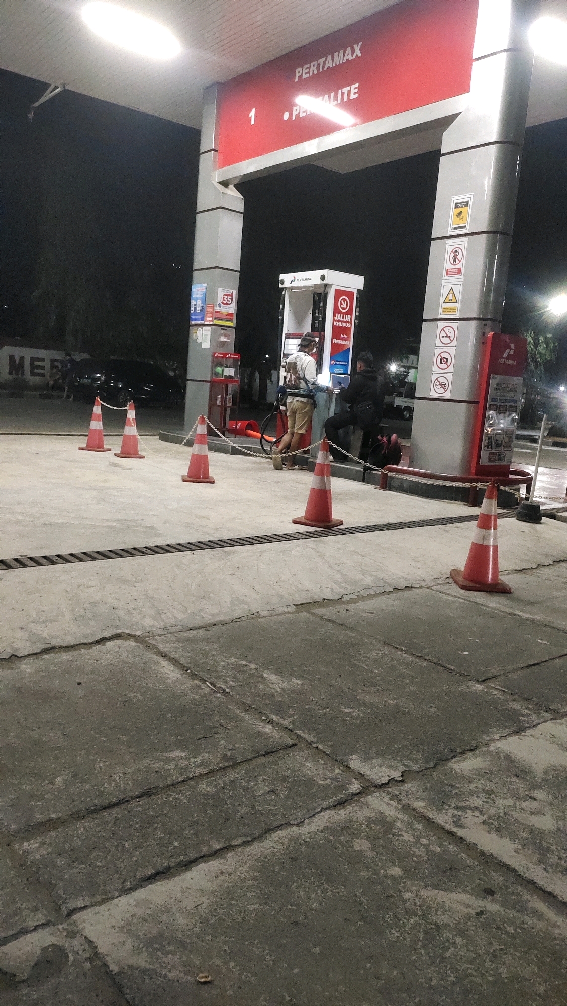 At a gas station in Menado, Indonesia, two individuals are present. One person, wearing a light-colored shirt and khaki shorts, is standing by a gas pump with a hose extended, appearing to be in the process of refueling. The other individual, dressed in dark clothing, is seated on a red bench near the pump, facing away from the camera. Several orange traffic cones are placed on the concrete ground, some connected by a chain, marking off areas. The gas station canopy is illuminated, with signage visible including "PERTAMAX" and "PERTALITE". Other signs with symbols and text are mounted on the pillars. The scene takes place at night.