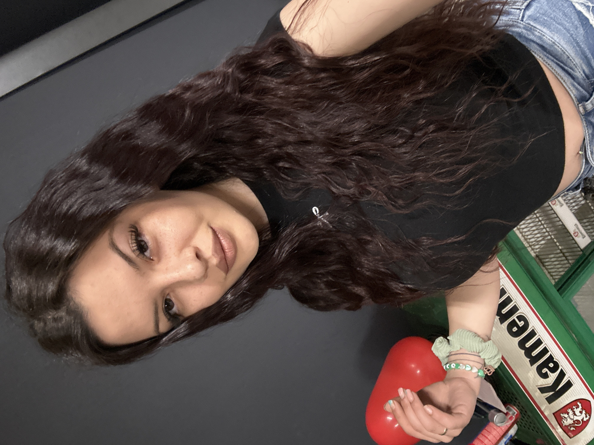 A young woman with long, dark, wavy hair is pictured from a high angle, seemingly taking a selfie. She has fair skin, dark eyes, and is wearing a black cropped top and blue denim jeans. Her midriff is visible. She is wearing bracelets on her left wrist and holding a red object in her hand. The background includes a dark gray surface and a green shopping cart with the word "KAMEN" in white letters along with a coat of arms. The location is presumed to be in the city of Sliven, Bulgaria, based on the brand "KAMEN," which is a local beer.