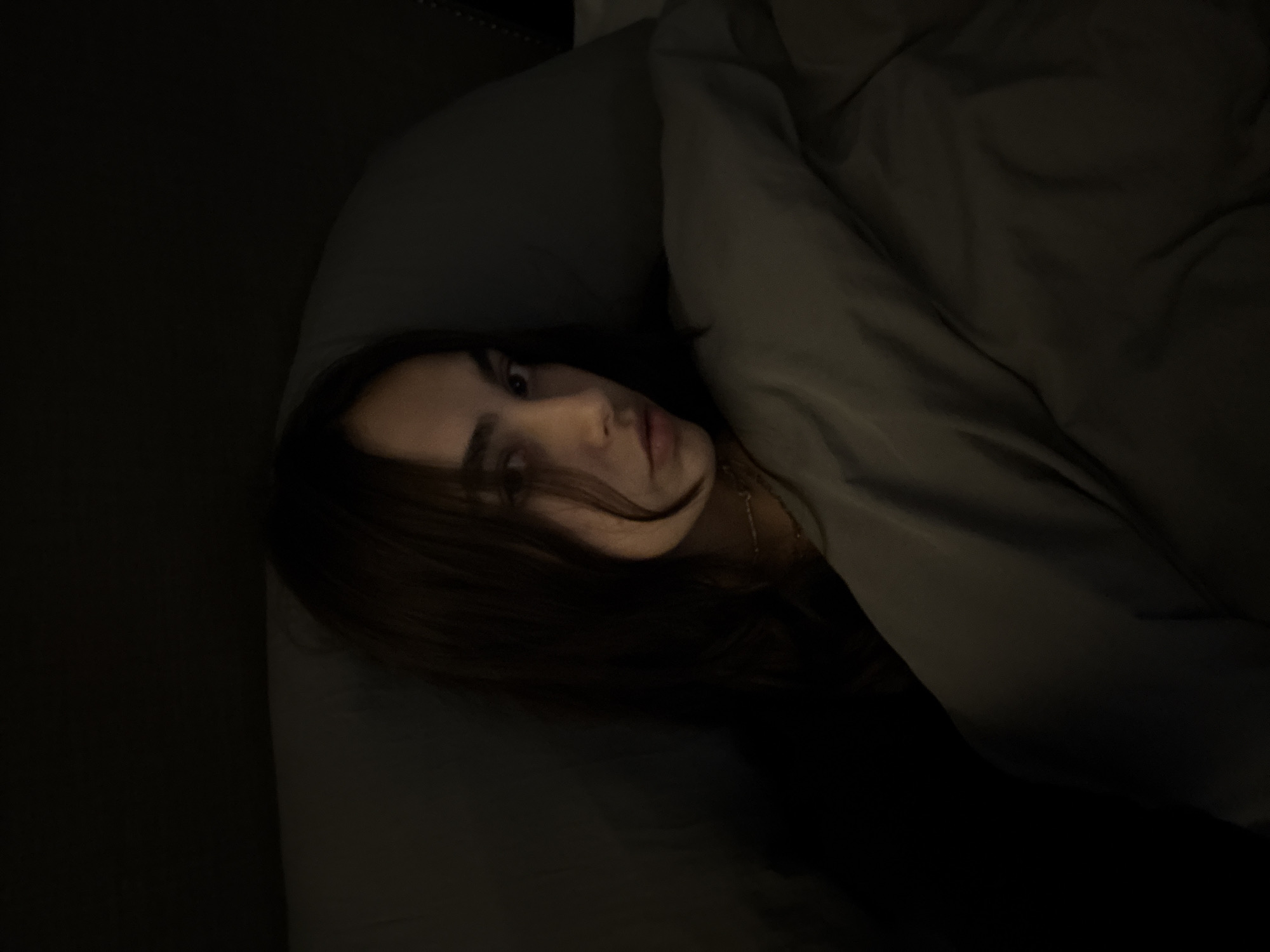 This photograph features a person, likely a young adult, lying in bed within a dimly lit room. The subject's face, with dark hair cascading around it, is partially illuminated by a soft light source, emphasizing one eye, the nose, and lips, as they gaze directly towards the camera. They are resting on a dark-colored pillow, with a matching dark grey or black blanket or duvet covering part of their body. A subtle glint of a thin necklace is visible around their neck.
The extreme low light conditions suggest the image was taken at night or in a very dark setting, creating a serene and intimate atmosphere. The person's expression appears neutral, possibly thoughtful or sleepy. While external context indicates the location is Mexico City, Mexico, there are no specific visual cues within the image itself to confirm this. No text is visible in the photograph.