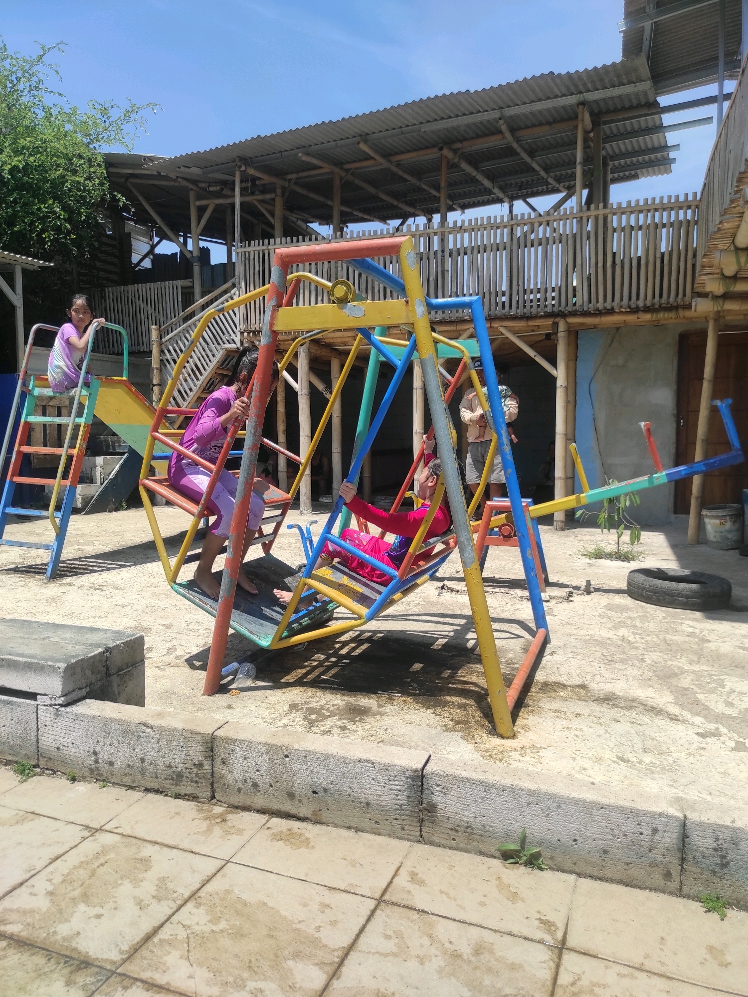 The image depicts an outdoor playground at a resort in Ngamprah, Indonesia, on a sunny day. A multi-colored metal swing set, yellow, red, blue, and green, is the central focus. Three children are on the swing set. A girl in a purple shirt and leggings is seated on the swing. Another girl in a pink shirt and dark pants is on the slide. A third child, wearing a pink shirt and dark pants, is on the other side of the swing set. In the background, a two-story structure with a corrugated metal roof and bamboo supports is visible. A wooden fence encloses part of the area. The ground is paved with concrete tiles and concrete blocks. A used tire lies on the ground near the swing set.