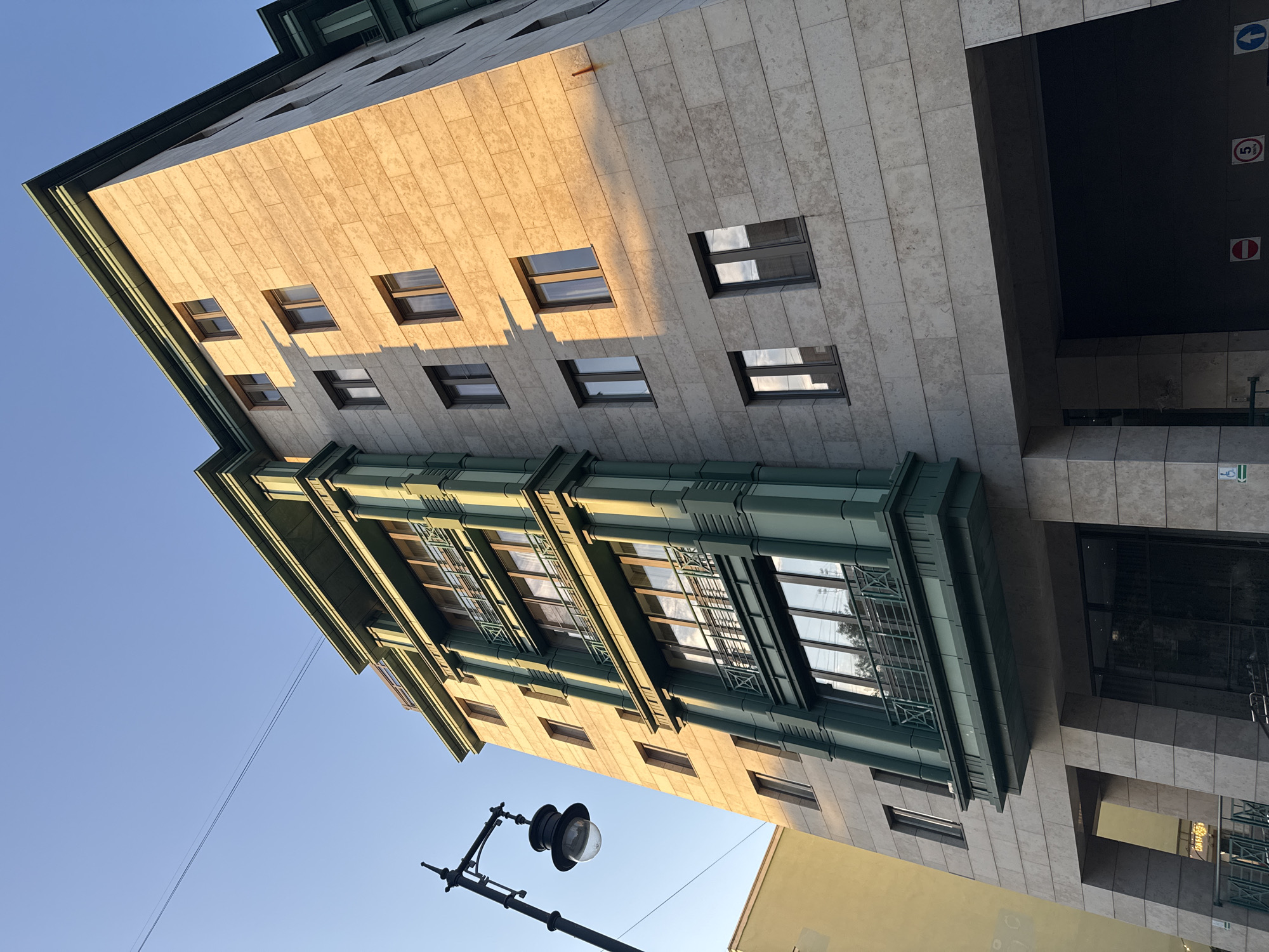 The image shows the facade of a modern building with a light-colored stone exterior and dark green accents on the balconies and window frames. The building has multiple stories, with rows of windows on each level. The sun is shining, casting long shadows on the building's surface. In the foreground, there is a black lamppost with an ornate lantern. Adjacent to the building, a portion of a yellow building is visible. The sky above is clear and blue. The building appears to be located in an urban setting, possibly a city, given the presence of the lamppost and the style of the architecture. While no people are visible in the frame, the scene conveys a sense of quiet urban life during daylight. The building's design suggests it might be residential or commercial, with balconies indicating individual units. The lighting suggests it's either early morning or late afternoon.