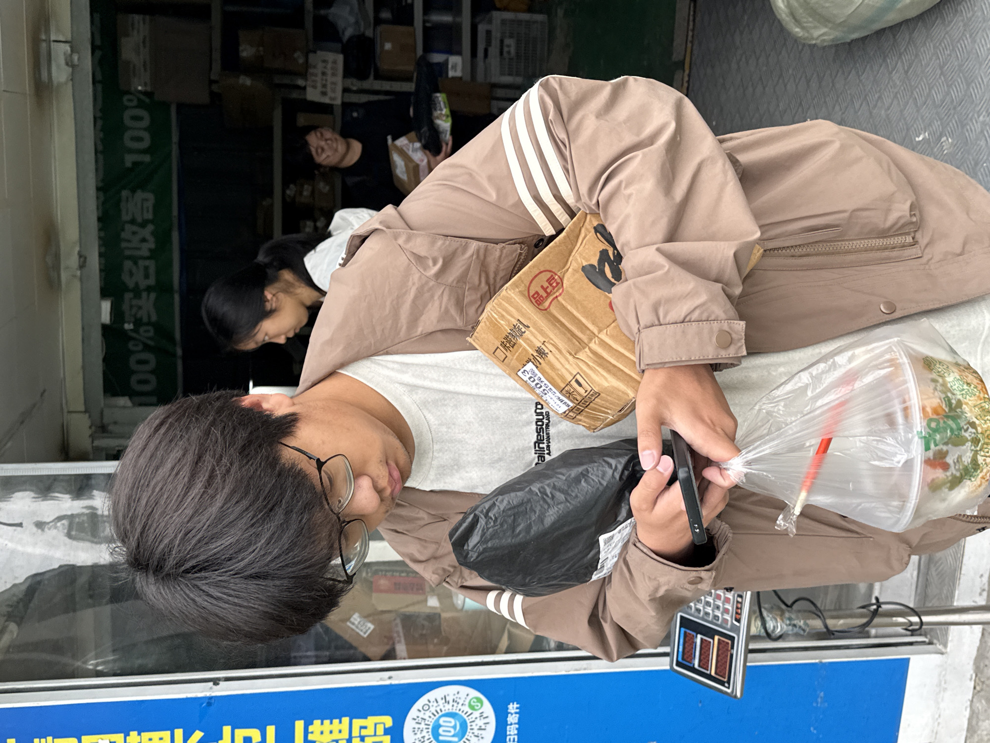 A man wearing glasses and a brown jacket stands outside a storefront in Jiangjin, China. He is looking down at his phone. He is holding a cardboard box, a black plastic bag, and a clear plastic bag containing food. Behind him, two other people are visible inside the store, surrounded by boxes. A blue sign with Chinese characters and a QR code is visible at the bottom of the frame. A weighing scale is also visible in the frame.