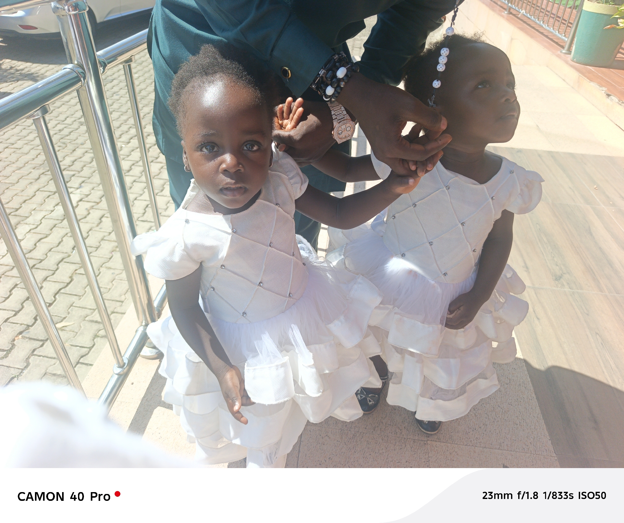Two young girls, likely twins, are dressed in matching white dresses with ruffles and embellishments. A person wearing a green outfit, a watch, and bracelets is holding their hands. They are standing near a silver railing on a paved surface. The photo was taken in the city of Barakin Lerawi, Nigeria.