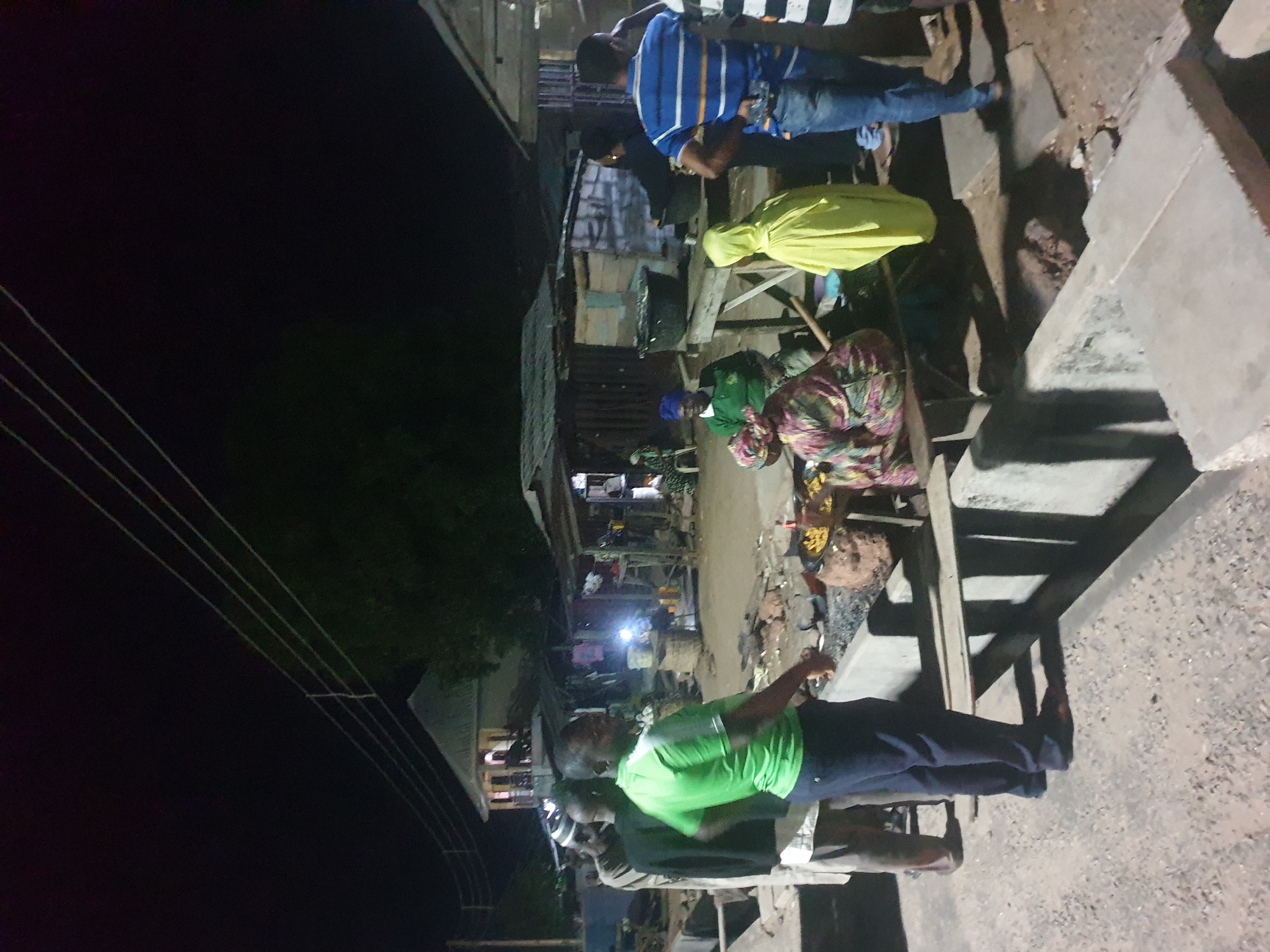 In Zango-ndaji, Nigeria, a nighttime street scene shows several people gathered. One man wears a green shirt and dark pants, standing near another person whose back is visible. Further down the street, a person is dressed in a yellow hooded garment, and another in a patterned robe or blanket. A man in a blue striped shirt is also present. The setting suggests an urban area with dwellings and structures visible along the street, illuminated by artificial light sources. Power lines stretch across the top left of the frame.