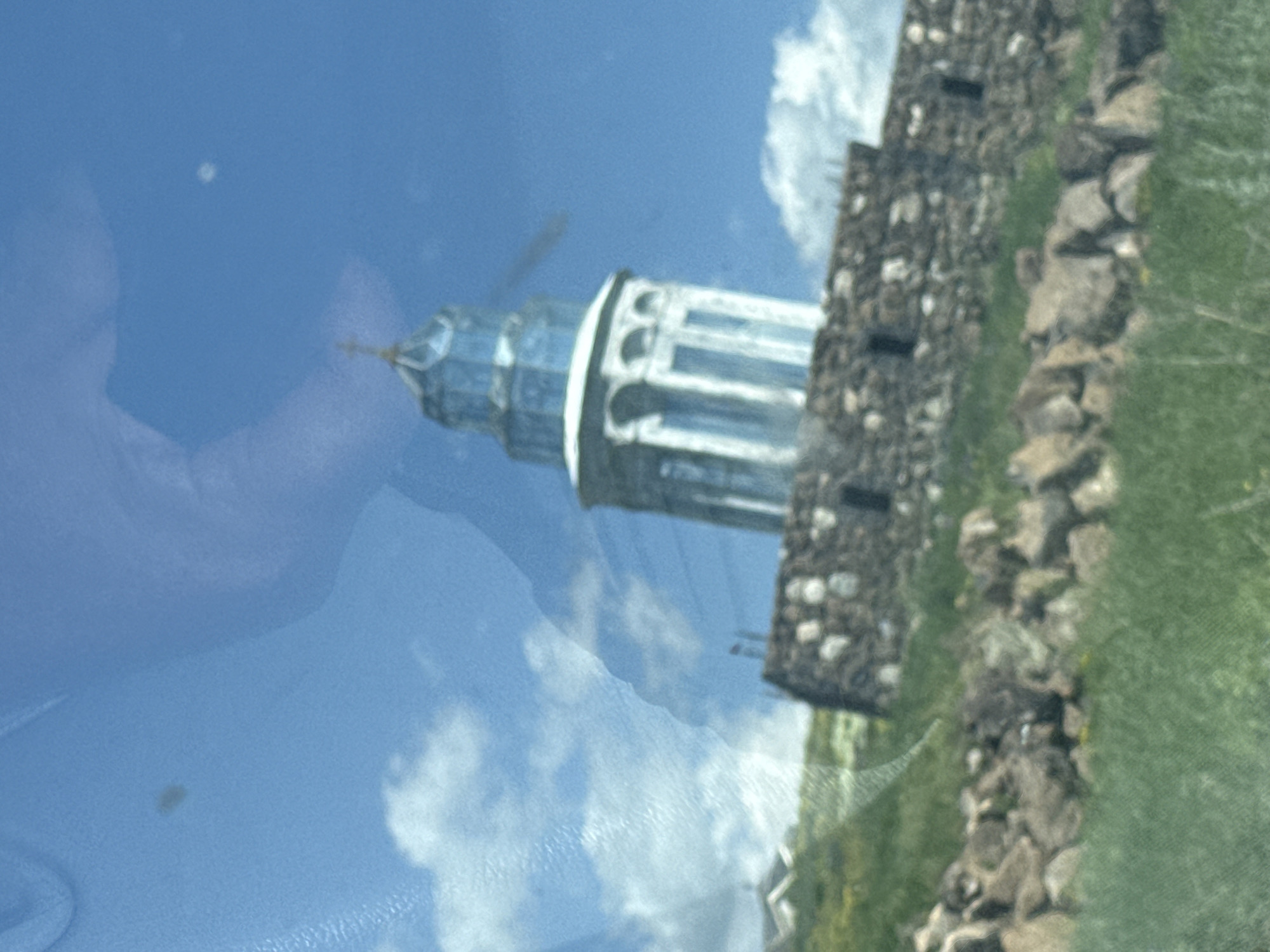 The image shows a lighthouse on a rocky hill with a bright blue sky and some clouds. There are reflections on the surface, obscuring parts of the image. The lighthouse has a white base with arched windows and a blue, glass-like top. The hill is covered with grass and scattered rocks. The image appears to be taken from a moving vehicle. This is likely the lighthouse in Artashavan, Armenia, situated on a hill overlooking a body of water, possibly Lake Sevan. The reflection in the foreground is unclear, but it could be a person or the interior of a vehicle. There are no clear interactions or actions visible due to the reflections and blurriness.