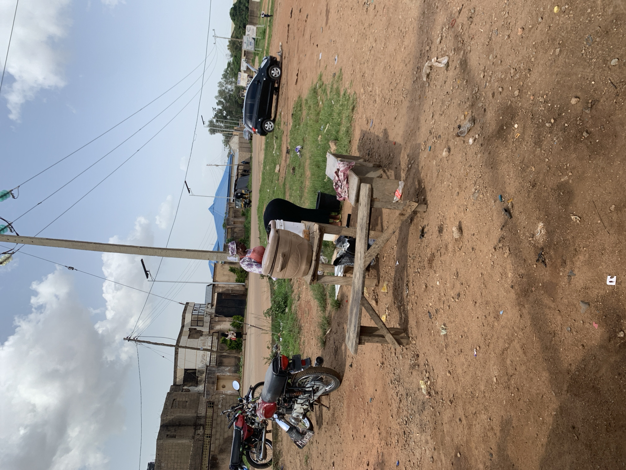 The image captures an outdoor scene in Barakin Lerawi, Nigeria, on a bright, cloudy day. In the foreground, a rustic wooden bench sits on reddish-brown, dusty ground littered with debris including plastic bottles and paper fragments. To the left, two motorcycles are parked; one red, and another slightly behind it, partially obscured, appears to be dark red or maroon. A woman, wearing a patterned headscarf and a red garment, is bent over a large, light brown clay pot positioned on a wooden stand, which also holds other items. Next to her, another person, dressed in black, is also engaged in an activity near the pot. In the middle ground, a black hatchback car is parked on a patch of green grass. In the background, several buildings are visible, some appearing unfinished, with a blue-roofed structure further back. Power lines crisscross the sky, supported by utility poles. The overall environment suggests a casual, possibly commercial, roadside setting.