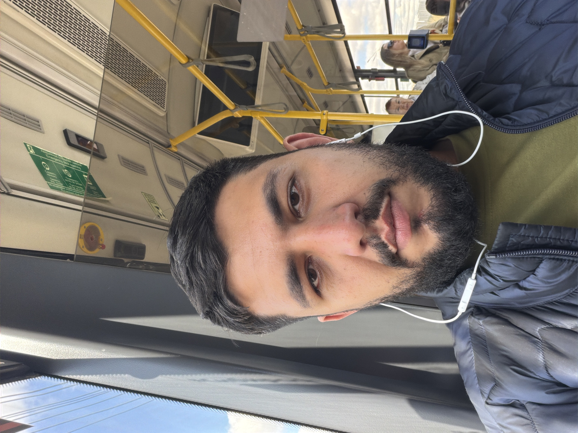 A man with a beard and headphones is riding on public transportation. Visible above and behind him are yellow handrails and the interior of a bus or train. Other passengers can be seen in the background. The scene is observed through a window, suggesting an urban setting. The specific location within Krasnoyarsk, Russia cannot be determined from the image.