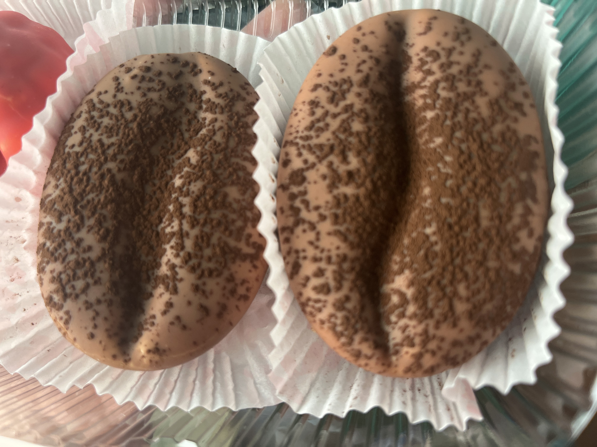 The image shows two chocolate confections, shaped like coffee beans, dusted with cocoa powder. They are presented in white, ruffled paper wrappers, suggestive of individual pastry packaging. The confections are arranged vertically, one above the other, with a slight overlap. The lighting is bright, highlighting the textures of the chocolate and the dusting of cocoa. The background is blurred, but appears to be a transparent container, possibly for displaying baked goods. There is a hint of red in the upper right corner, possibly another sweet or fruit. The location in Desyatikhatka, Russia, is not evident from the visual information provided in the image.
