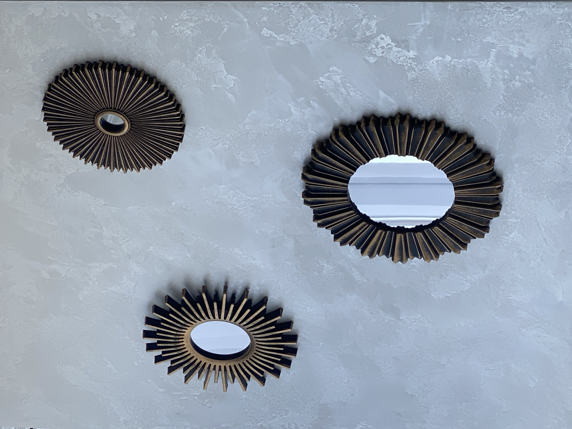 Three sunburst-style decorative mirrors with ornate bronze frames are mounted on a textured gray wall. The mirrors are oval in shape and vary slightly in size and the intensity of their sunburst pattern. The wall has a rough, stucco-like finish with variations in light and shadow. The image is taken from a medium distance, showing the arrangement of the mirrors on the wall. There is no indication of any people or specific location details within the image itself.