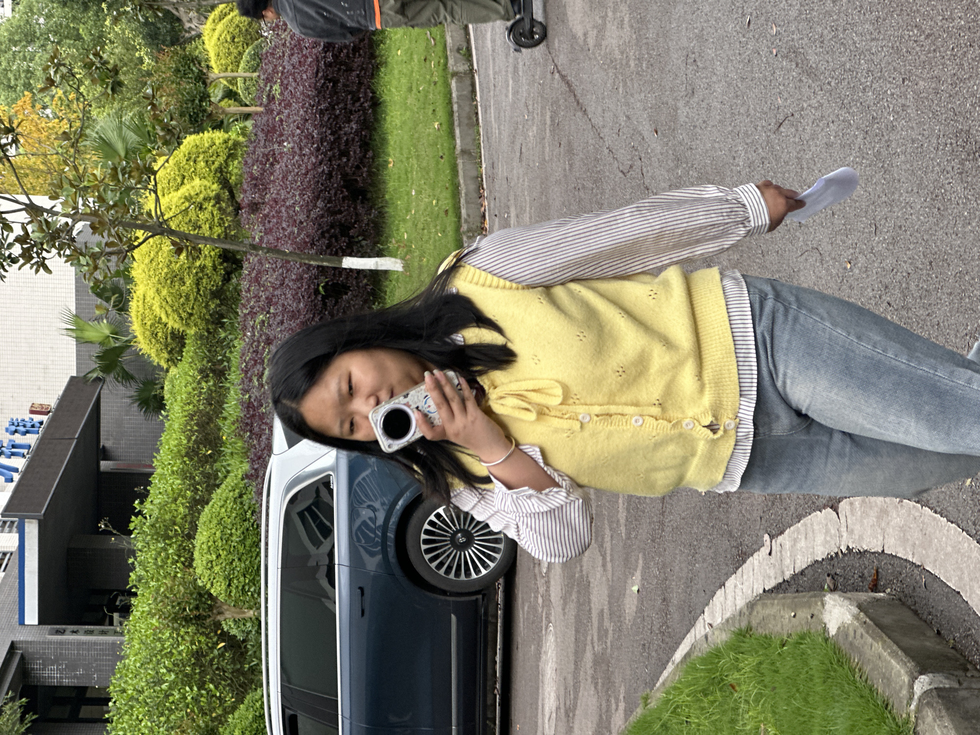 A young person, likely female, is captured standing outdoors on a paved surface, looking down at a white and silver cylindrical device held to her face, possibly a compact camera or similar gadget. She wears a light yellow knitted vest over a striped long-sleeved shirt and blue jeans, while holding a small crumpled piece of white paper in her right hand. A dark blue car is parked behind her, with its front passenger wheel and side visible.
The scene is rich with greenery, including bright green rounded bushes, darker foliage, and reddish-purple hedges. Trees with slender trunks and dark leaves are also scattered throughout. In the far background, a modern building with dark grey/black walls and blue trim is visible. Faint Chinese characters on the building, consistent with the Jiangjin, China context, appear to spell out "Yangtze Normal University." Another individual, partially visible in the upper right, is seen from behind, pushing a wheeled object along the paved path.
The lighting is natural and diffused, suggesting an overcast day. The overall setting is an outdoor, well-maintained area, possibly a university campus or park.