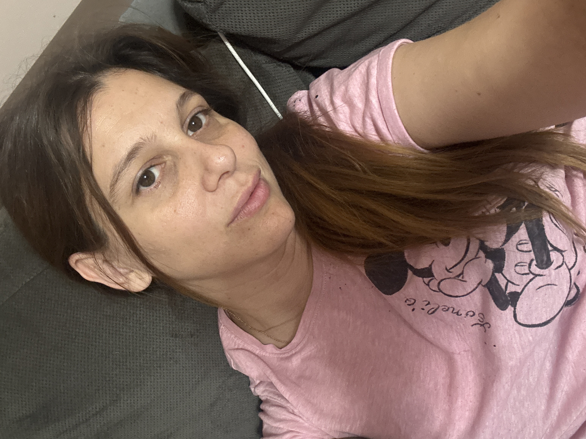 A young woman with light brown hair is lying down on a gray sofa, taking a selfie. She is wearing a pink t-shirt with a Mickey Mouse graphic. The woman is looking directly at the camera with a neutral expression. The lighting is natural and appears to be coming from the side. The background is out of focus, but it seems to be an indoor setting. There is no information in the image to indicate the location in Plovdiv, Bulgaria.