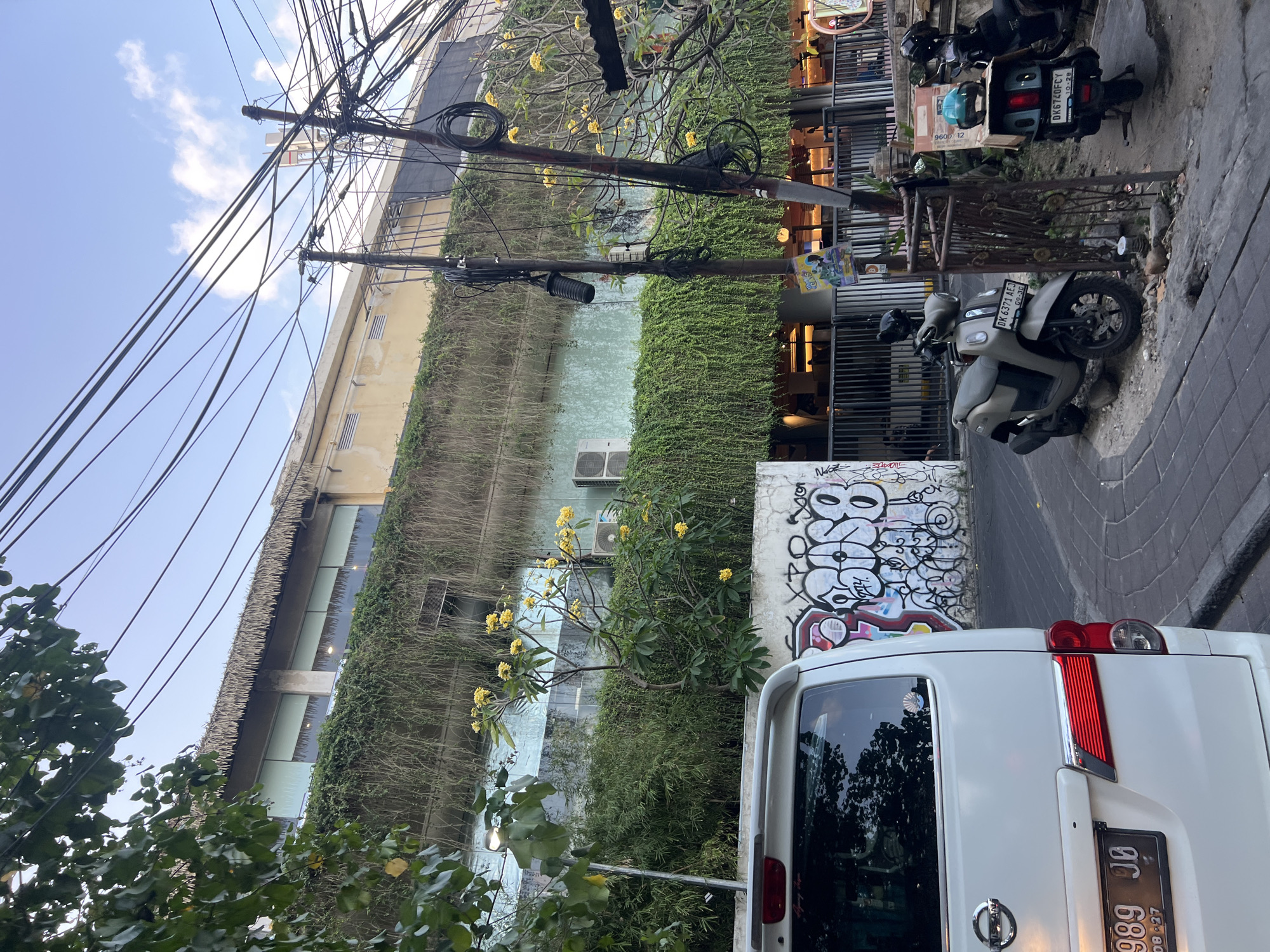 In Kuta, Indonesia, a white van is parked on a street. The van is facing away from the camera and is on the left side of the image. The van has a license plate that reads "B 989 JO". To the right of the van, there are two scooters parked. The scooter on the left is a light brown color and has a license plate that reads "DK 6371 AEJ". The scooter on the right is dark blue and has a license plate that reads "DK6740FCY". Behind the van and scooters, there is a building with a thatched roof and greenery growing on its facade. The building has windows and air conditioning units. There are also electrical wires and poles in the background. The street is paved with bricks. Graffiti is visible on a wall to the left of the van.