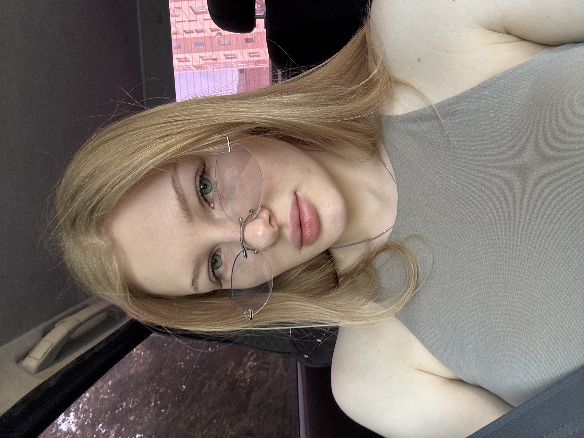 A young, fair-skinned woman with blonde hair and green eyes is pictured in what appears to be the interior of a car. She is wearing rimless glasses and a gray top. Her body is angled toward the right, but her face is turned towards the camera. A building is visible through the car window. The location of the image is unknown; no indicators suggest the city of Perm, Russia.