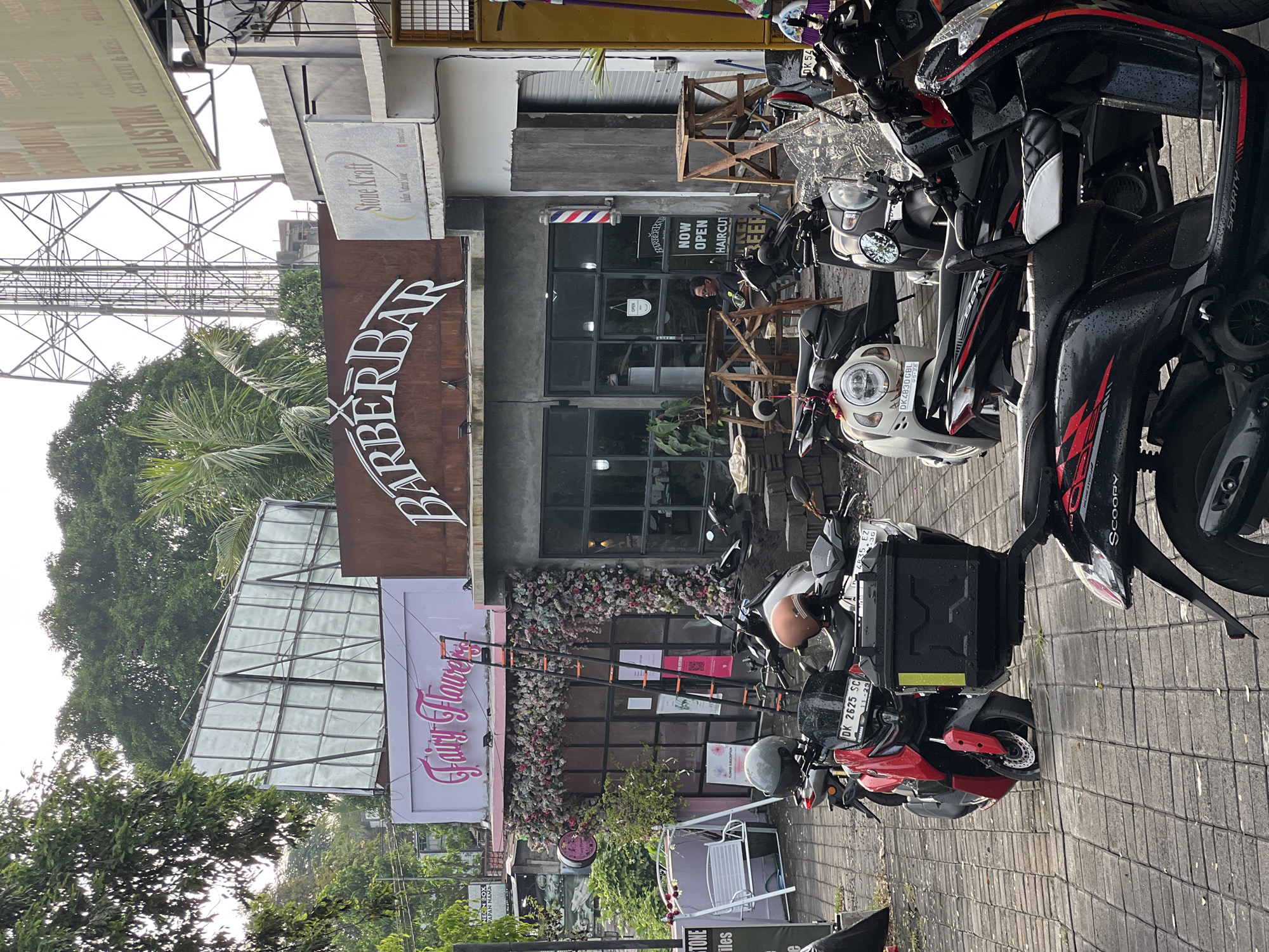 This image captures a vibrant street scene in North Kuta, Indonesia, featuring several commercial establishments and parked motorcycles. The scene is dominated by shopfronts along a paved sidewalk, with lush green trees and an overcast sky in the background.
The most prominent businesses visible are a "BARBERBAR" with a large, rustic brown sign and a classic barbershop pole outside its glass-fronted entrance. A "NOW OPEN HAIRCUT" sign is displayed on its window, and a person is faintly visible inside. Next door is "Fairy Flowers," distinguished by a pink sign with flowing script and extensive floral decorations cascading around its windows. Above the barbershop, another sign reads "Stone Kraft." Further down the street, a larger sign is partially visible, indicating "ALAT LISTRIK," suggesting an electrical tools shop.
Parked along the sidewalk are several scooters and motorcycles, typical transportation in the region. Notable models include a Honda Scoopy and another with "SPORTY" branding. License plates such as "DK 2625 SC," "DK 4830 GBL," and "DK 54" are visible on the vehicles. The pavement appears slightly damp, possibly from recent rain. Various wooden structures, perhaps for outdoor seating or temporary construction, are also present near the shops. The overall impression is one of a bustling commercial area.
