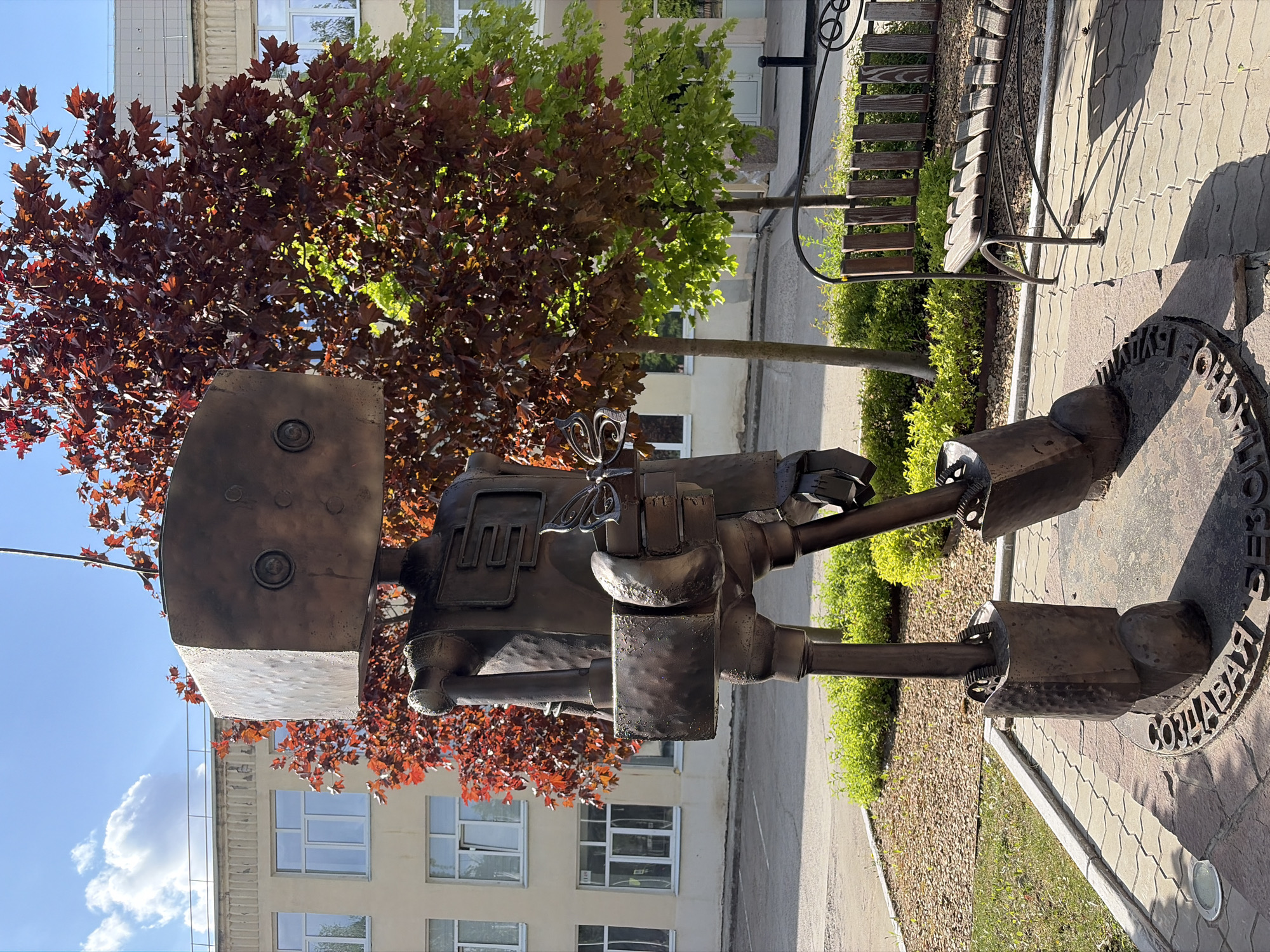 The image shows a robot statue in the city of Chaikovsky, Russia. The bronze-colored robot has a boxy head with two circular eyes. It has arms and legs made of pipes and gears. The statue is placed on a circular pedestal with writing around the edge. Behind the statue, there is a building with windows and a tree with both red and green leaves. To the side of the statue, there is a bench with a dark metal frame and wooden planks, and a paved walkway. The sky is blue in the upper left corner.