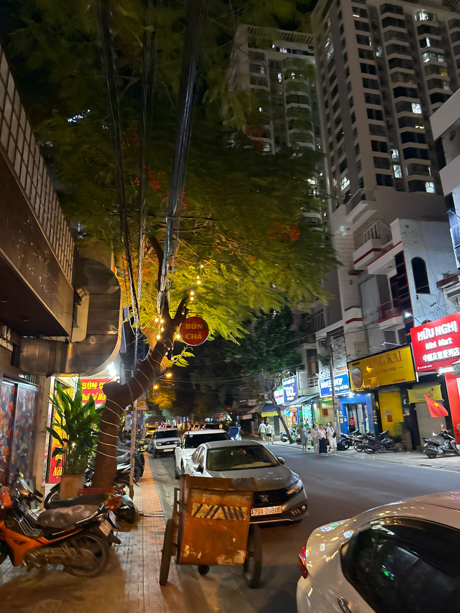 The image depicts a street scene at night in Nha Trang, Vietnam. The street is lined with multistory buildings, some of which are illuminated. A large tree with vibrant green foliage and red flowers is prominent in the foreground, decorated with a string of lights. Several vehicles, including motorcycles and cars, are parked along the street or are in motion. A cart with an orange frame and weathered wooden planks is visible on the sidewalk. Signs in Vietnamese are displayed on the buildings, indicating businesses. People are seen walking along the sidewalk and across the street. The overall atmosphere suggests a busy urban environment at night.