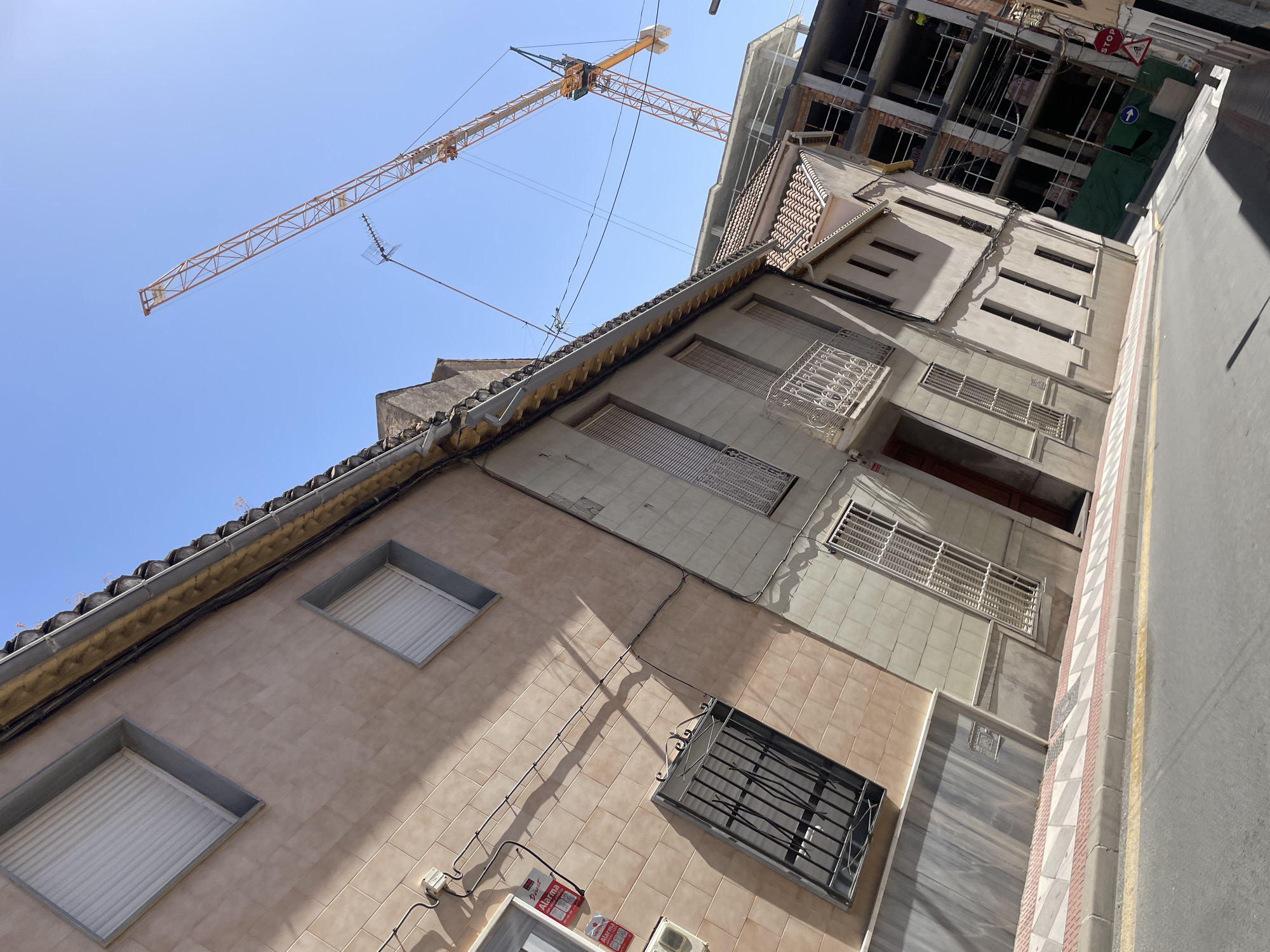 The image depicts a street scene in Atarfe, Spain, on a clear, sunny day. The perspective is from street level, looking up at buildings and a construction site. On the left, a two-story building with a pale pink tiled facade features two windows with white roller shutters. Electrical conduits and an alarm box are visible on the wall. Below one window is a black metal security grate. The sidewalk in front of this building has a patterned tile border.
Adjacent to this building, on the right, is a narrower, taller building with a light beige facade, also featuring windows with roller shutters and a decorative white metal balcony. Further to the right, a multi-story building is under construction, with exposed brickwork, concrete floors, and scaffolding. A large orange construction crane towers over the scene, its boom extending across the bright blue sky. In the background, a few power lines and an antenna are also visible. The street itself is paved asphalt with a yellow line marking the edge and a patterned pedestrian walkway. Traffic signs are visible on the right side of the street, including a stop sign and a sign indicating a bicycle route. No people are clearly visible in the image, though a construction worker in a yellow vest may be present on one of the upper floors of the construction site. The overall impression is one of urban development and ongoing construction.