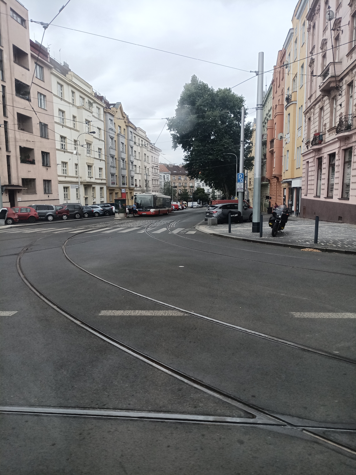 The image depicts a street scene in Prague, Czechia, featuring a tram track junction in the foreground. Several multi-story buildings with colorful facades line the street on both sides. On the right side of the street, a black motorcycle is parked on the sidewalk. Further down the street, a red and white tram is stopped at a crosswalk. Several cars are parked along the left side of the street. Overhead, tram wires crisscross the sky, and a large tree with dark green foliage is visible behind the buildings on the left. The sky is overcast, suggesting cloudy weather. The image appears to be taken from a moving tram, indicated by the perspective and the tram tracks filling the lower portion of the frame.
