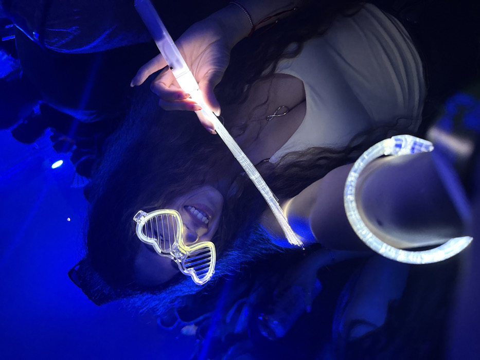 An overhead-angled, close-up photograph captures a smiling female subject with long, wavy dark hair, illuminated by blue ambient light and personal glowing accessories. The scene occurs within an indoor entertainment venue, likely a nightclub or concert hall, located in Miami, United States of America. The precise address or name of the venue is not visually ascertainable from the image.
The subject wears translucent, heart-shaped novelty glasses with horizontal slats, which emit a soft yellow glow on her face. She is dressed in a white tank top, revealing a silver-colored infinity symbol pendant necklace. A thin red string bracelet is visible on her right wrist. Her left arm is extended towards the camera, prominently displaying a glowing, translucent, C-shaped bracelet adorned with a serpent motif. Her right hand securely holds a long, translucent fiber optic wand, which emits bright white light from its base and along its fibers. The background features indistinct, silhouetted figures of other individuals, consistent with a crowded social event under deep blue illumination. The woman exhibits a broad smile, directly facing the camera.