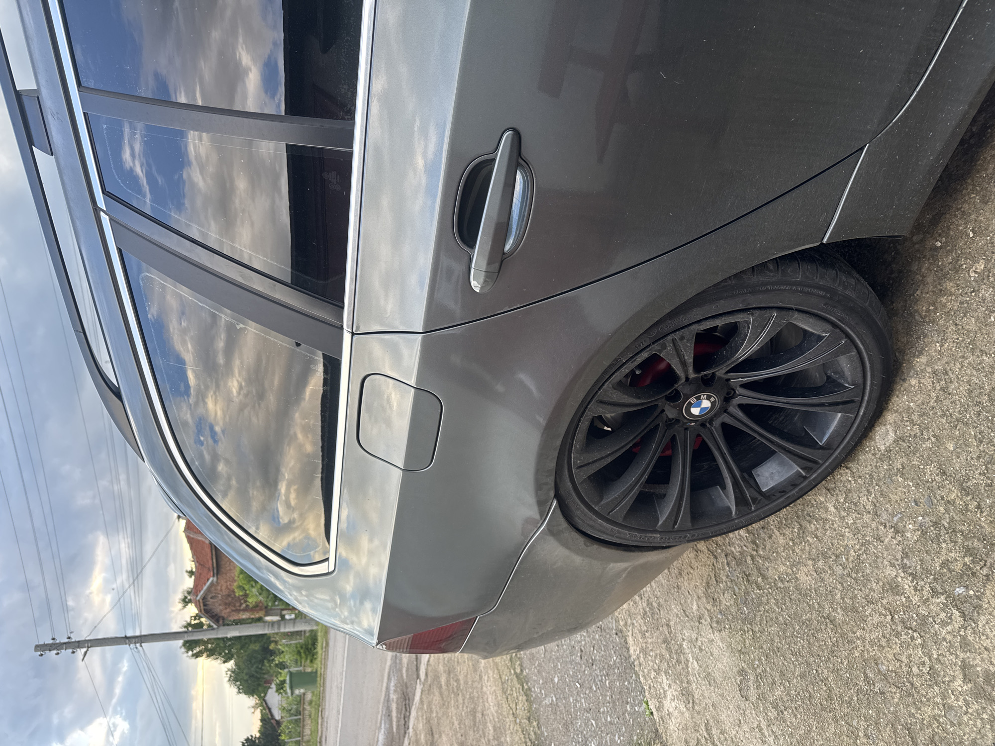 An image, rotated 90 degrees counter-clockwise, captures the rear passenger side of a dark grey BMW touring car (likely a 5-series, E61 generation) parked outdoors in Sarnevo, Bulgaria. No people are visible.
The car, appearing somewhat dusty, is equipped with black multi-spoke alloy wheels, featuring a prominent BMW logo on the center cap. Red brake calipers are discernible behind the spokes. The tires, identified as Pirelli P Zero, display text including "RADIAL TUBELESS," "XL," "MAX. LOAD," "MAX. PRESS.," "TREADWEAR 300," "TRACTION AA," "TEMPERATURE A," "E4," "0202973," and "DOT" followed by numbers. The windows are tinted, reflecting a partly cloudy sky with warm, soft light, suggesting late afternoon or early evening. Chrome trim lines the window frames and roof rails. A fuel cap is visible on the rear fender.
The background, seen through and reflected on the car's windows, reveals a rural or suburban setting. Reflections show a building with a terracotta roof, green foliage, utility poles, and overhead power lines. Small regulatory markings like "E4 43R-010356," "AS3," "Transp. % L 26," "23 DOT," and "ECE R43" are visible on the car's windows. The car is parked on a rough concrete or asphalt surface.