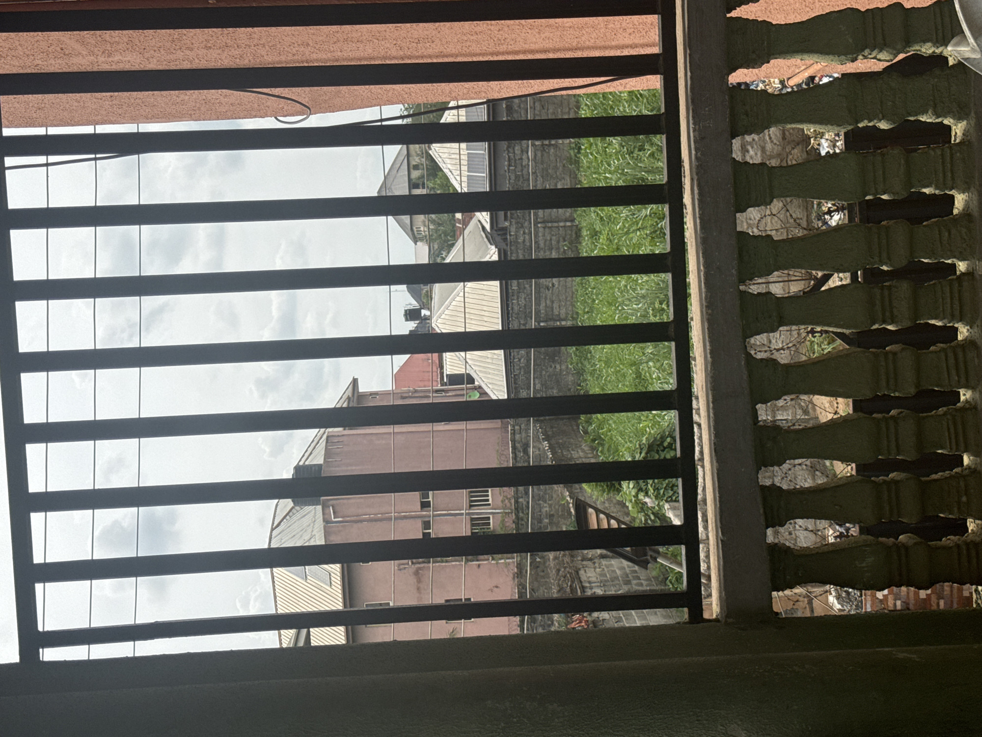 A view through a window security grill and decorative concrete block wall. Outside, there are buildings and greenery, suggesting an urban residential setting. The buildings are a mix of single and multi-story structures, with pink and grey facades. The image was most likely taken in Ihiagwa, Nigeria. No people are visible in the image.