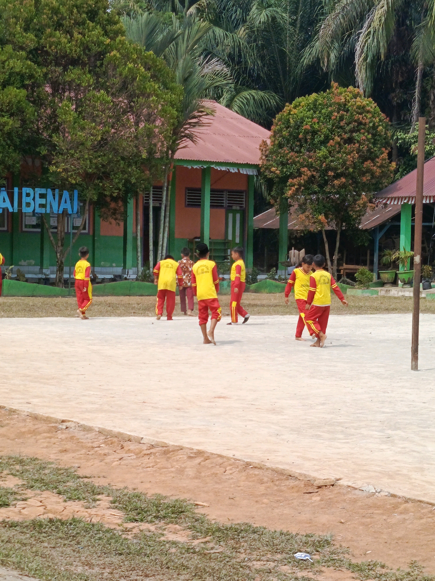 A group of children, likely students, wearing matching yellow shirts with red trim and red pants are playing on a dirt field in front of a school building. The school building appears to be painted orange and green, with a red roof. Trees and palm trees surround the school. The sign above the school entrance is partially visible and reads "AI BENAI". The overall scene depicts a typical school environment in a tropical region.