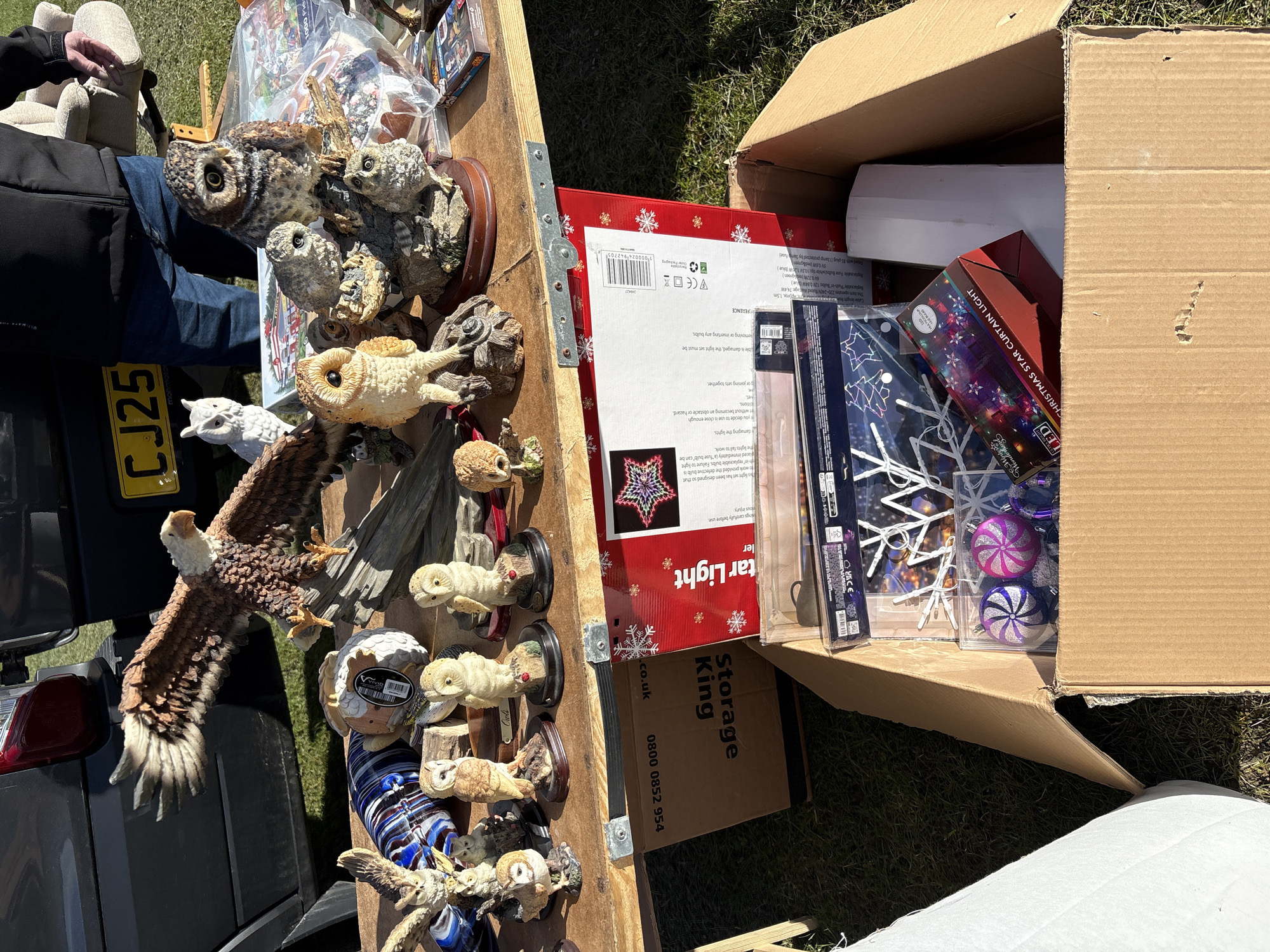 The image shows a collection of items for sale at what appears to be a car boot sale or outdoor market. The items are arranged on a wooden surface and inside a cardboard box. The items include several owl figurines, an eagle figurine, Christmas star curtain lights, Christmas ornaments, and other miscellaneous items. A vehicle, likely a car, is visible in the background, with a license plate that reads "CJ25". The location appears to be outdoors, with grass visible in some areas. Text "Storage King" and a phone number are visible on a box. The location is Chirk, United Kingdom.
