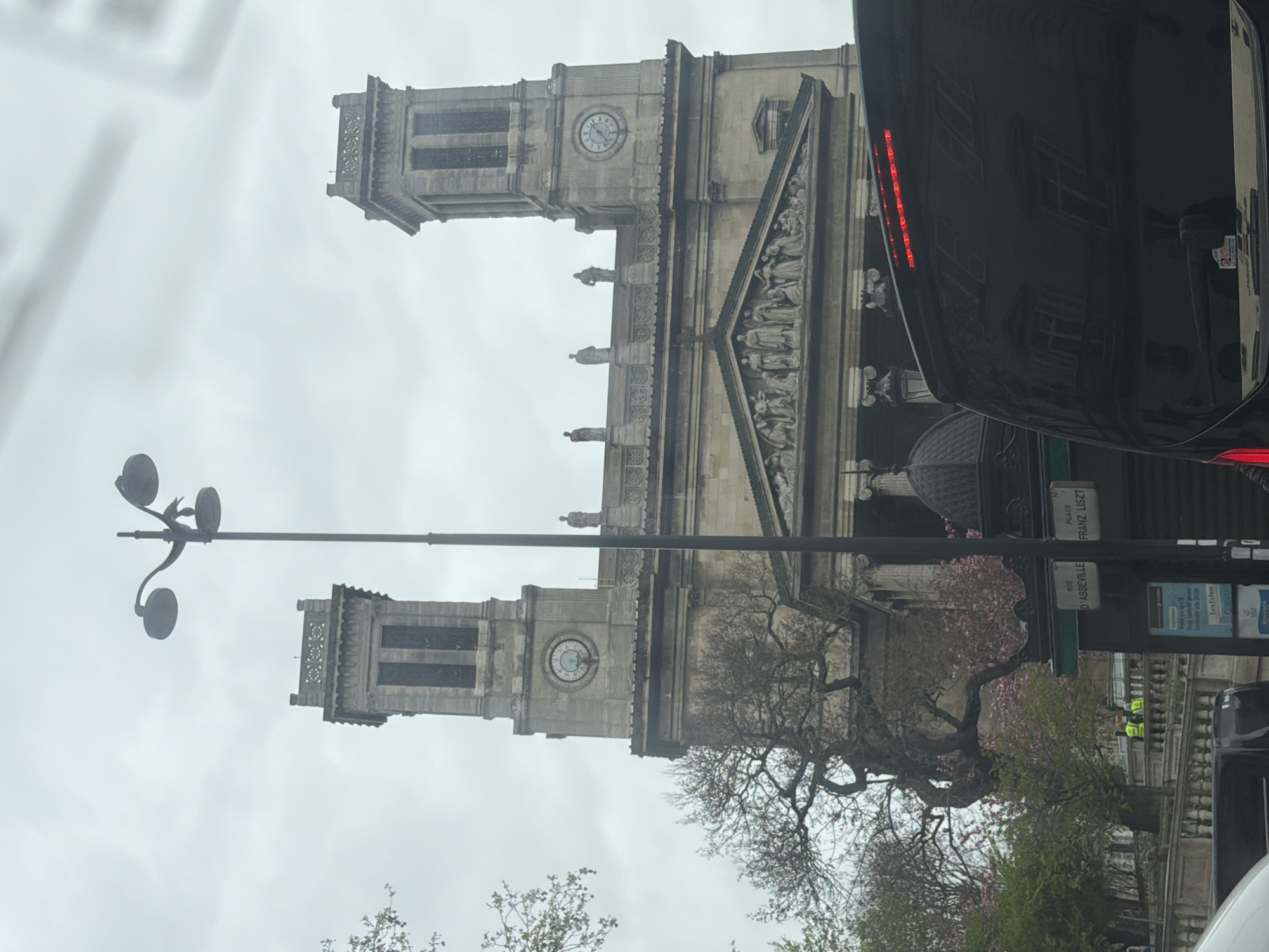 The image shows the Église Saint-Vincent-de-Paul in Paris, France. The church has two towers, each with a clock face. A lamppost is in front of the building. A portion of a black vehicle is visible in the foreground. The street sign reads "Place Franz Liszt" and "Rue d'Abbeville." Some trees with pink blossoms are also visible. Two individuals wearing yellow vests can be seen along the building's walkway. The sky is overcast.