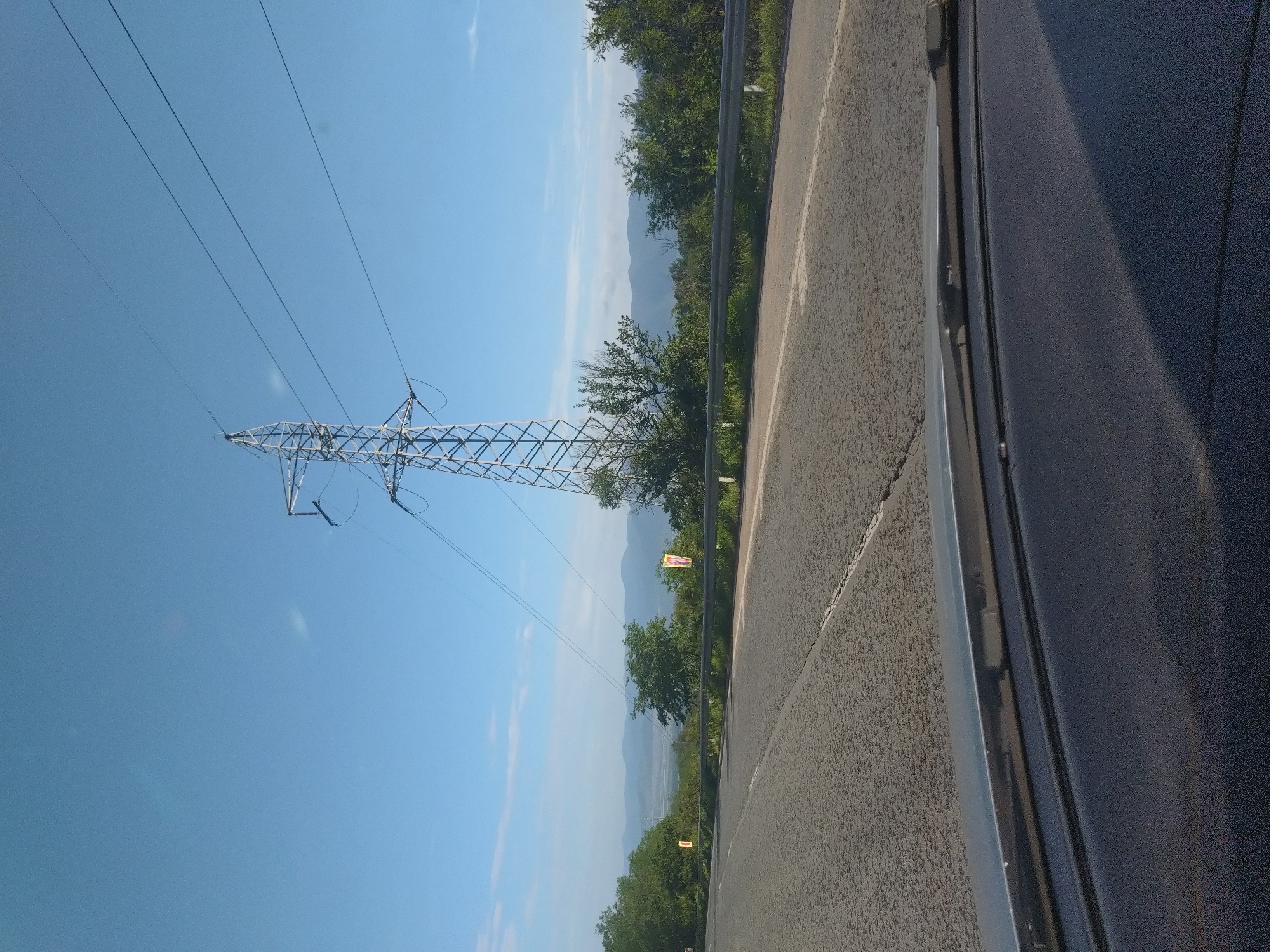 The image presents a daytime view from inside a vehicle, looking forward through a windshield, with a dashboard and a windshield wiper visible at the bottom. The scene depicts an asphalt road curving slightly to the left in a rural or semi-rural environment near Zmeyovo, Bulgaria.
On the right side of the road, a silver metal guardrail runs parallel to the asphalt. Beyond the guardrail, a dense line of green bushes and trees extends into the midground. A large, metallic lattice power transmission tower (pylon) stands prominently on the right, its structure reaching high into the clear, light blue sky. Multiple power lines extend from the top of the pylon, crossing horizontally across the upper right portion of the sky.
To the left of the road, more green trees and bushes are visible. In the distance, a range of undulating mountains or hills appears hazy against the horizon, indicative of a clear but somewhat distant view. The sky above is largely clear and light blue, with a few faint, wispy white clouds. Two vertical, rectangular banners or signs with indiscernible details are situated among the trees, one on the left side of the road and another further down on the right. The road itself appears to be a two-lane surface with a solid white line marking its right edge and a broken white line in the center, suggesting a two-way traffic flow. The asphalt surface exhibits a coarse texture with visible cracks and imperfections. No individuals or specific actions are discernible within the frame.