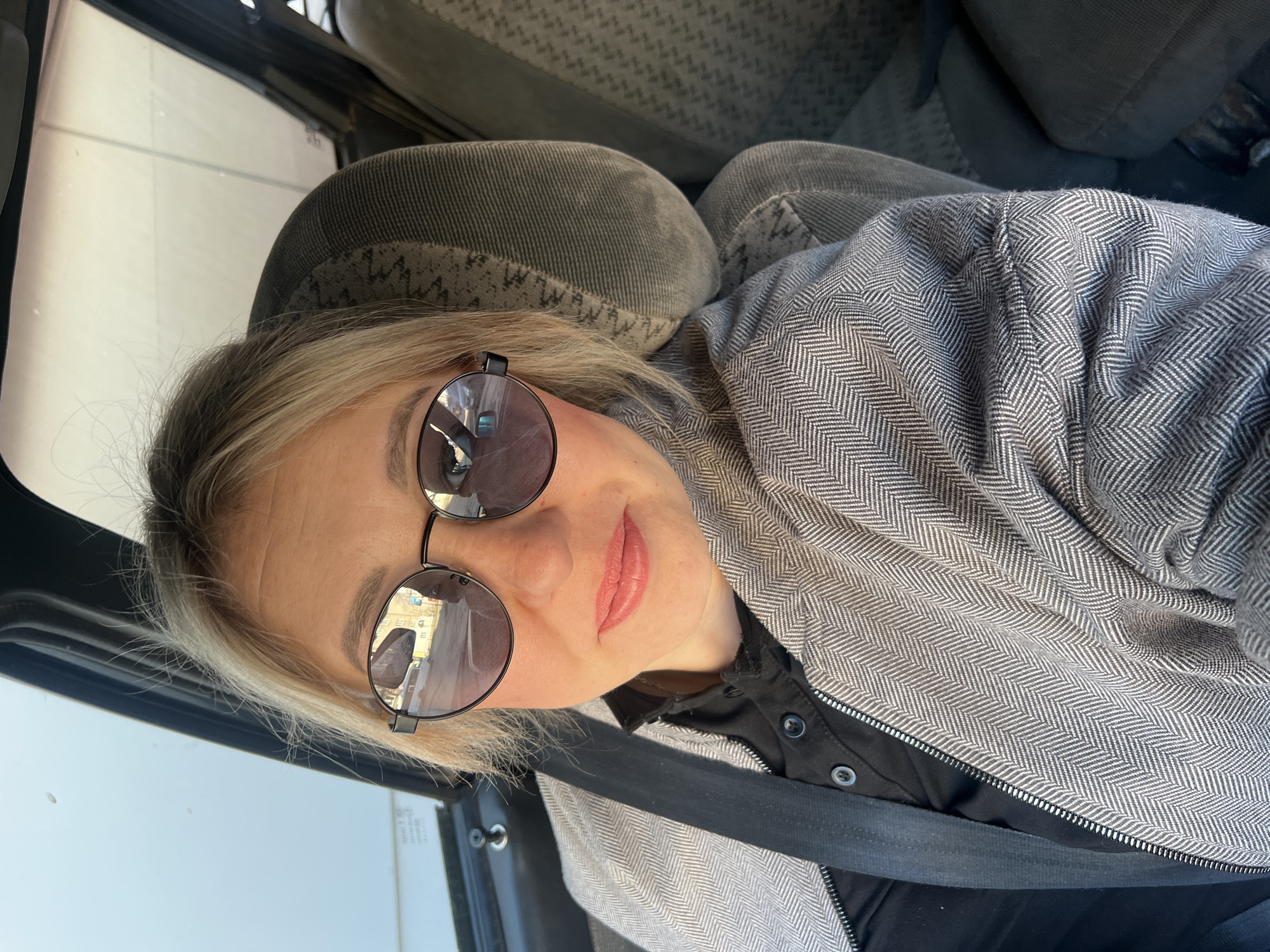 A sideways selfie captures a blonde-haired woman seated in the passenger seat of a car, set in Chaikovsky, Russia. She wears round, dark sunglasses that reflect a street scene with buildings and vehicles, indicating it is daytime. Her lips are adorned with light pink lipstick. She is dressed in a black collared shirt and a grey jacket featuring a prominent herringbone pattern. A black seatbelt is fastened across her body. The car's interior, including textured grey seats and a window with faint, unreadable text, is visible in the background. She looks directly at the camera with a subtle, pleasant smile.