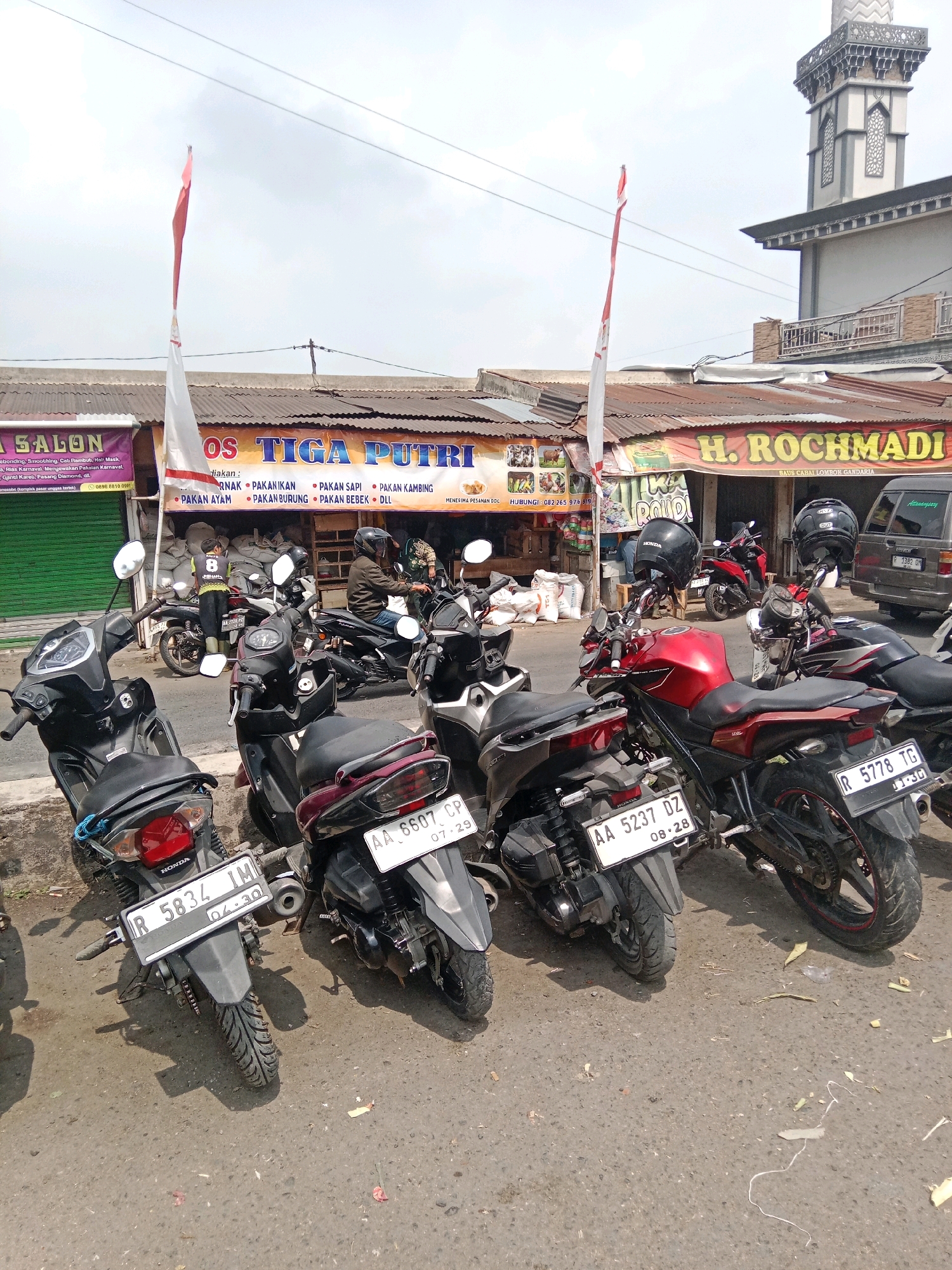The image shows a street scene in Kertek, Indonesia, likely near a commercial area. In the foreground, several parked motorcycles are visible, with their license plates indicating registration. One motorcycle in the lower left has the license plate R 5834 IMD. To its right, a black scooter has the license plate AA 6607 CP. Further to the right, a red motorcycle has the license plate AA 5237 DZ. Another black motorcycle in the background has the license plate R 5778 TG.
Behind the motorcycles, a row of shops lines the street. One shop displays the name "TIGA PUTRI" and lists various animal feed products such as "PAKAN ANAK," "PAKAN IKAN," "PAKAN AYAM," "PAKAN BEBEK," and "PAKAN KAMBING." Another shop is named "H. ROCHMADI." A red and white Indonesian flag is prominently displayed, suggesting a national holiday or celebration. A minaret of a mosque can be seen in the upper right portion of the image. Two individuals are visible on motorcycles; one person wearing a helmet and a yellow shirt with the number 8 on it is standing next to a motorcycle. Another person wearing a jacket and helmet is seated on a motorcycle. The environment appears to be an outdoor street with an overcast sky. The ground is paved with asphalt and shows signs of wear, with some debris and fallen leaves. The shops have corrugated metal roofs and simple facades.