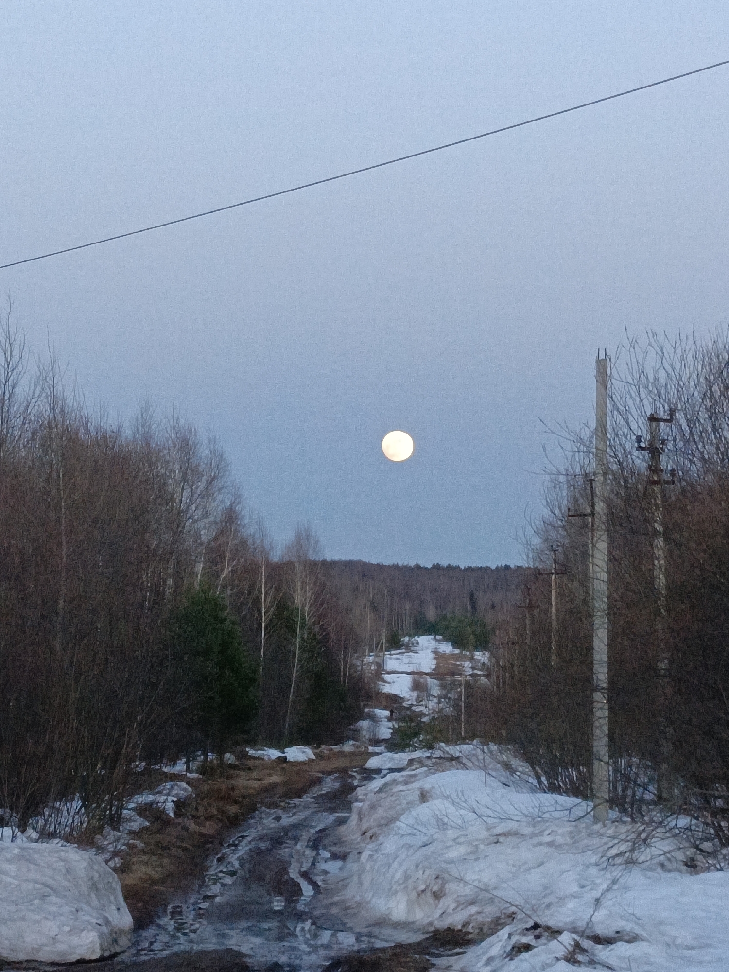 The image depicts a dirt road in a rural area, possibly on the outskirts of Марковский, Russia. Patches of snow remain on the ground and along the sides of the road, indicating early spring or late winter. The road is wet and muddy in places. On either side of the road are trees, mostly bare of leaves, suggesting a deciduous forest. A few evergreen trees are visible. Power lines run above the road, supported by utility poles. In the distance, a hill or elevated landform is covered in trees. In the sky, a full moon is prominently visible. No people are visible in the image. The overall lighting suggests either early morning or late evening.